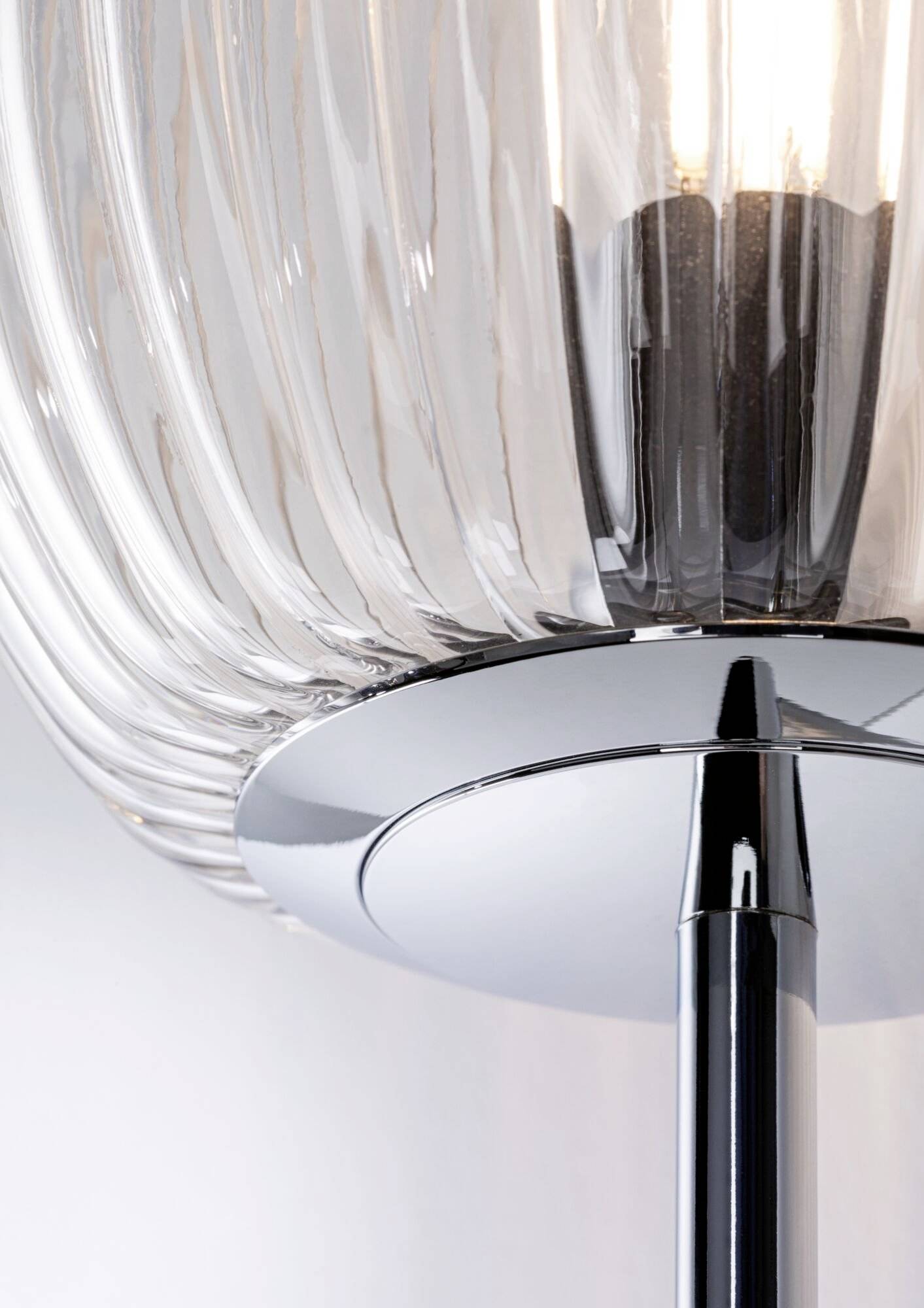 Detailní pohled na moderní stojací lampu s vroubkovaným sklem a chromovanou základnou, která vyzařuje jasné, teplé světlo.