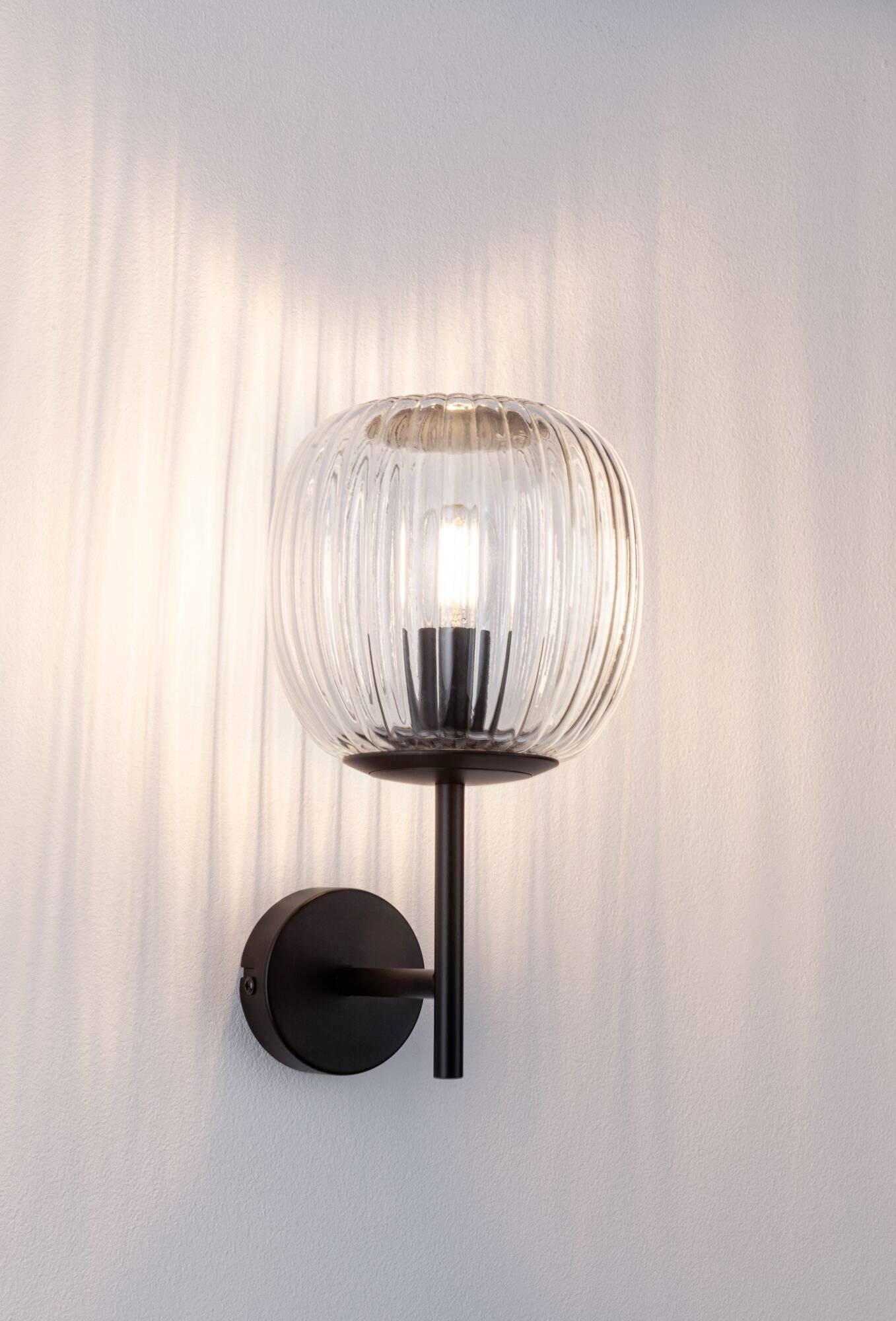 Nástěnná lampa s černým kovovým rámem a žebrovaným sklem obklopujícím žárovku. Světlo vzorně dopadá na stěnu.