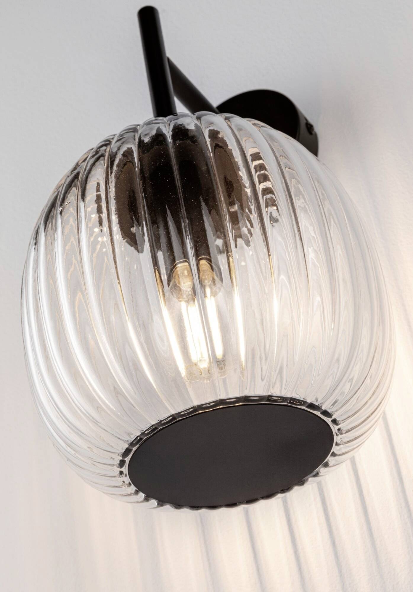 Nástěnná lampa s vroubkovanou, průsvitnou skleněnou koulí a černým držákem, která vyzařuje teplé světlo.