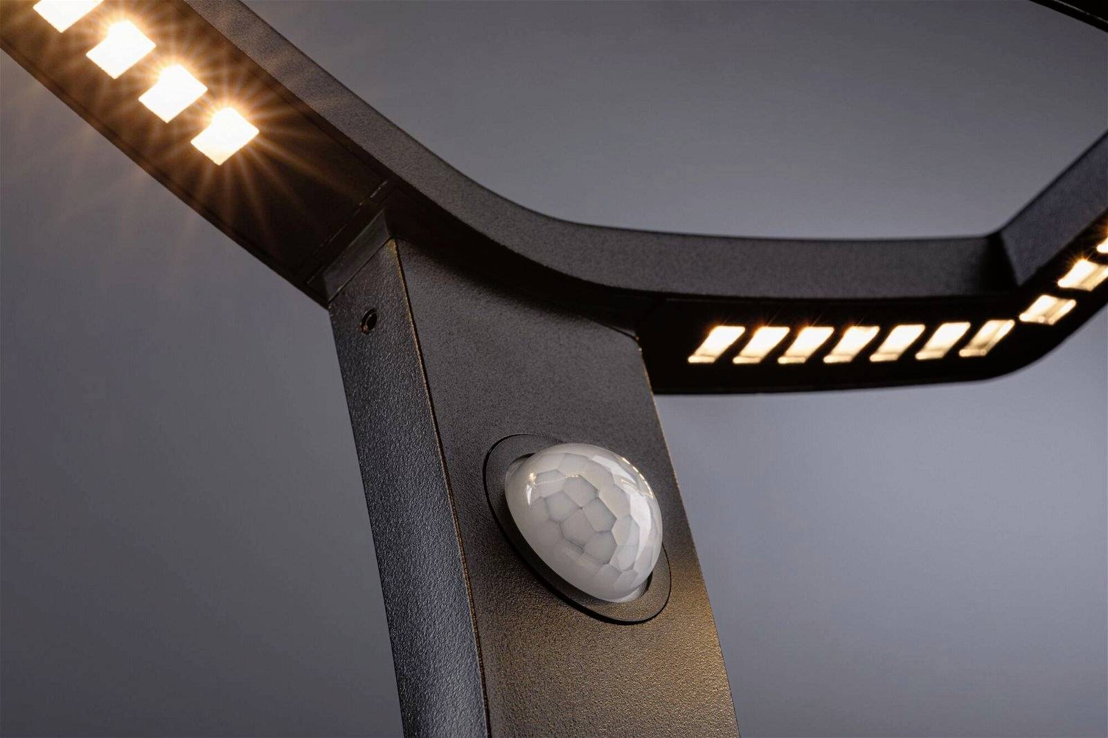 Detailní záběr černého kovového objektu s osvětlenými LED diodami a bílým senzorem uprostřed, před šedým pozadím.