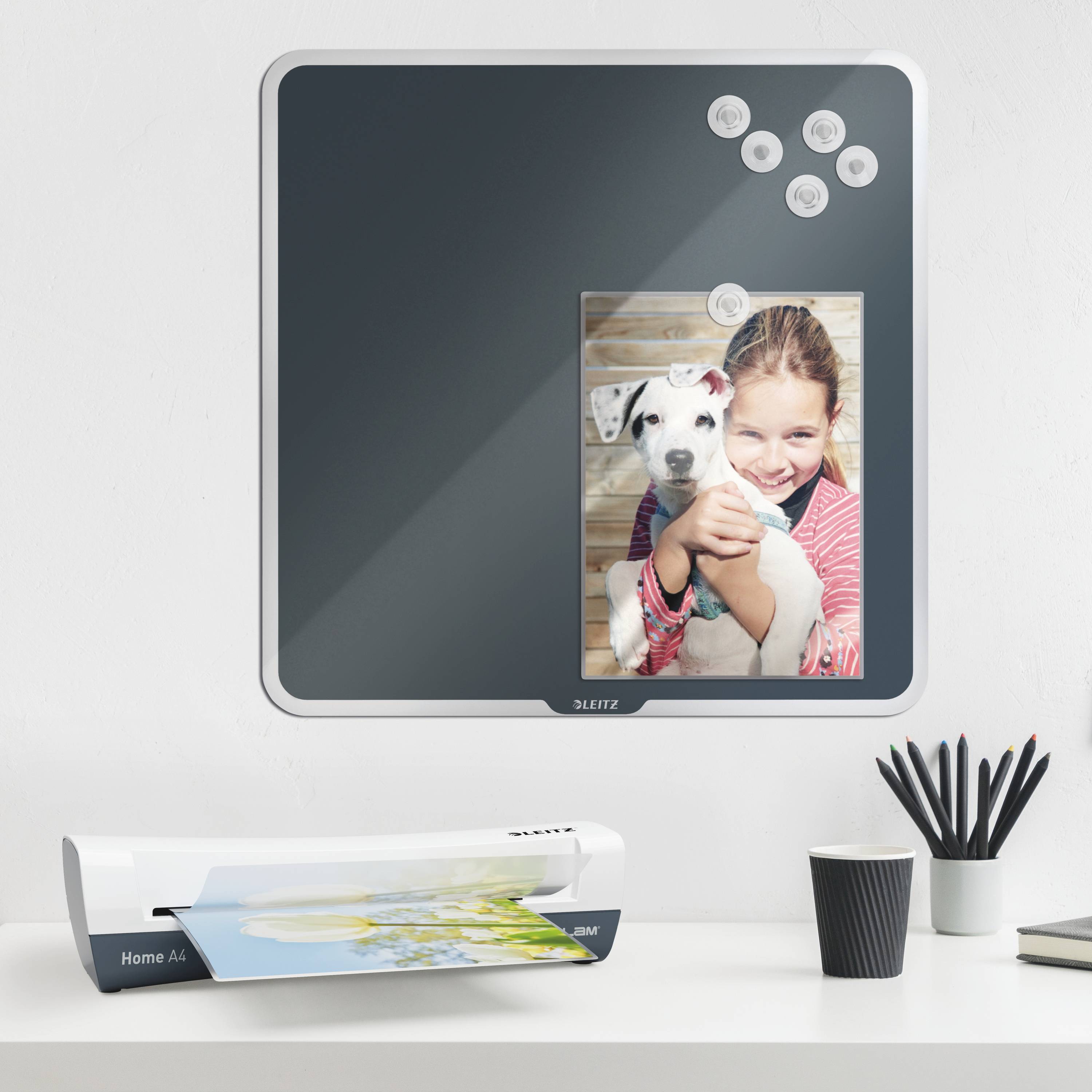 Magnetická tabule na stěně s fotografií dítěte objímajícího psa, vedle kancelářské potřeby na stole, včetně tiskárny a šálku.
