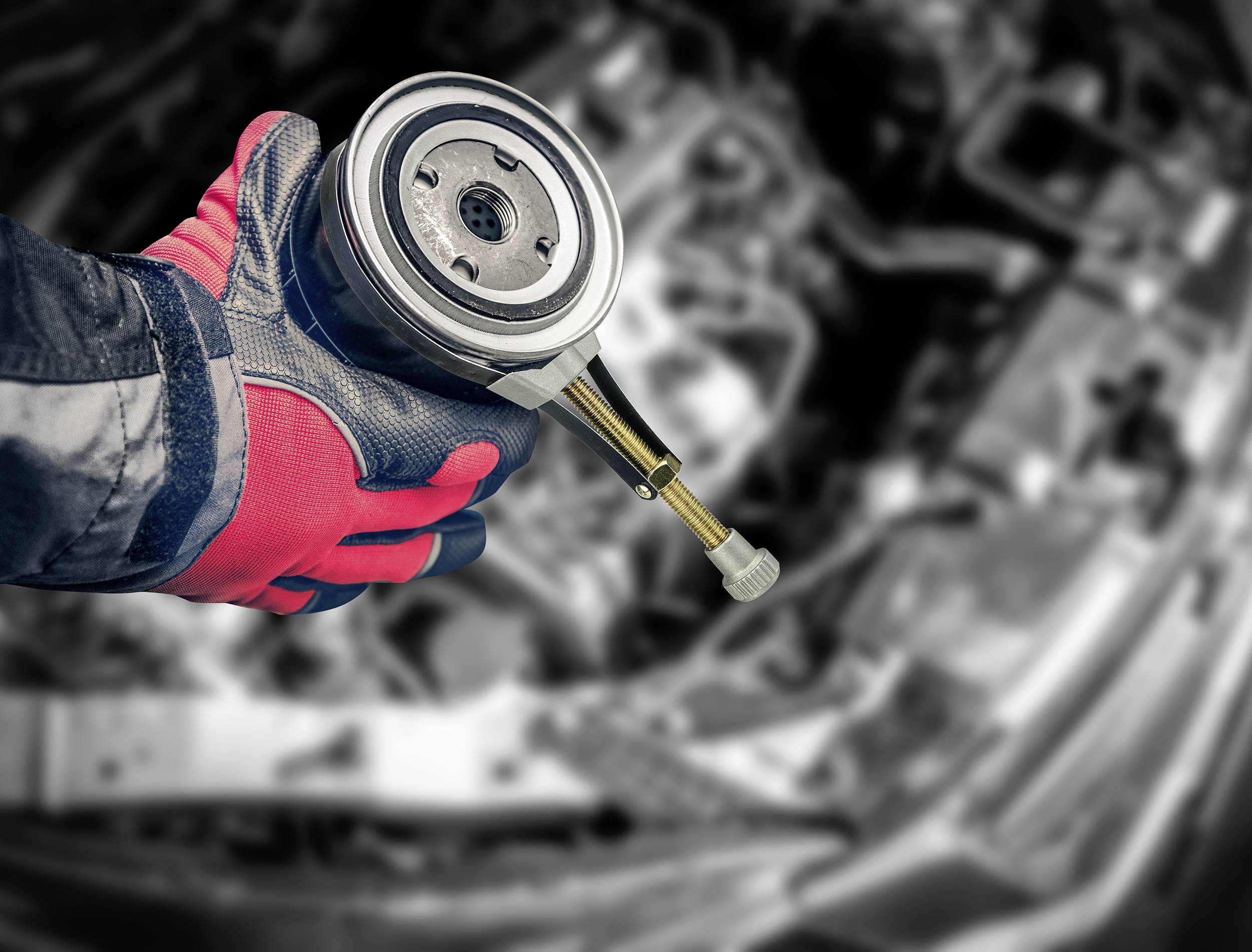 Rukavicí opatěná ruka drží automobilovou součástku před rozostřeným pozadím s otevřeným motorovým prostorem.