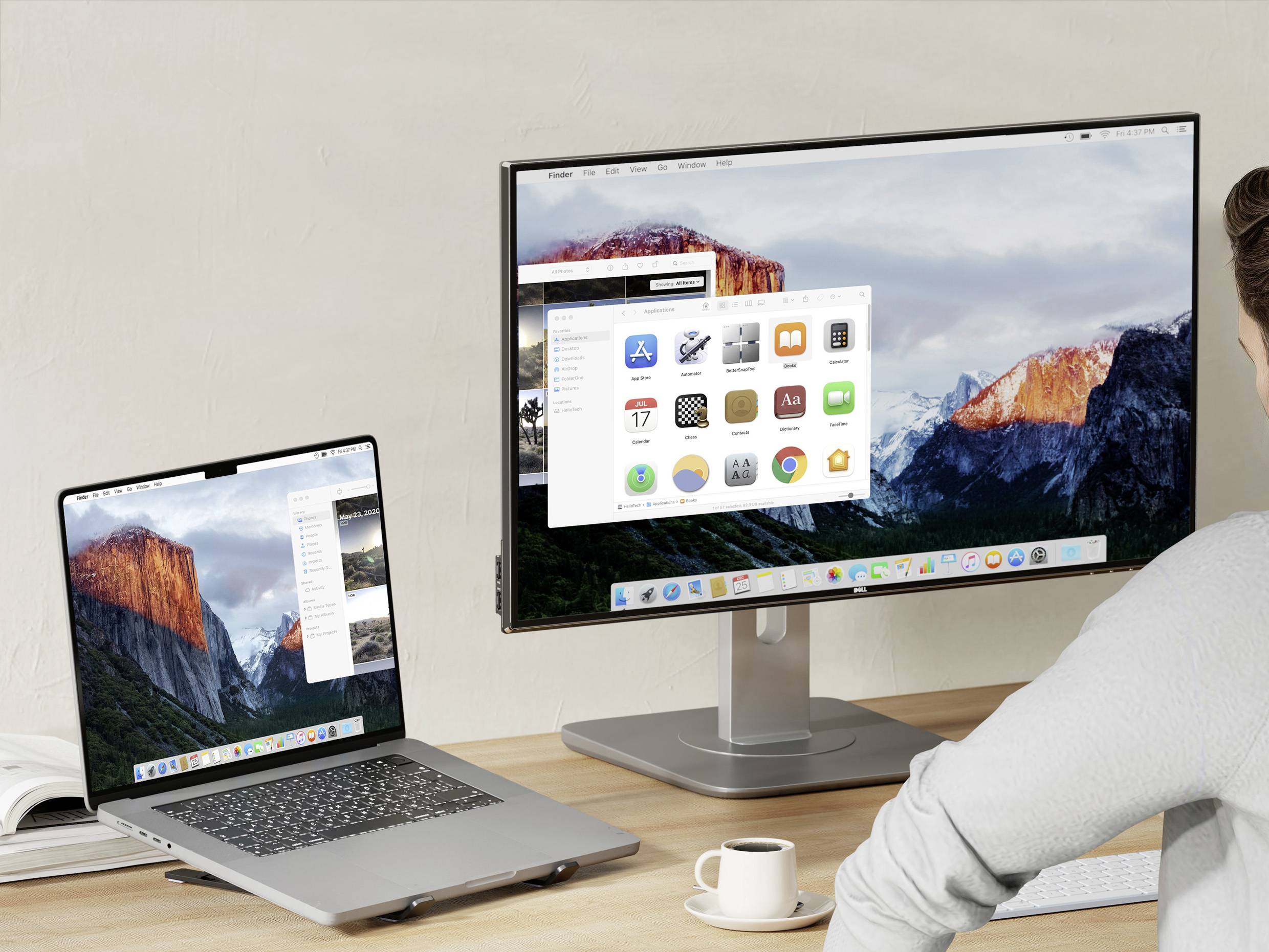 Osoba sedí u stolu s laptopem a monitorem zobrazujícím rozhraní macOS s aplikacemi a krajinným pozadím.