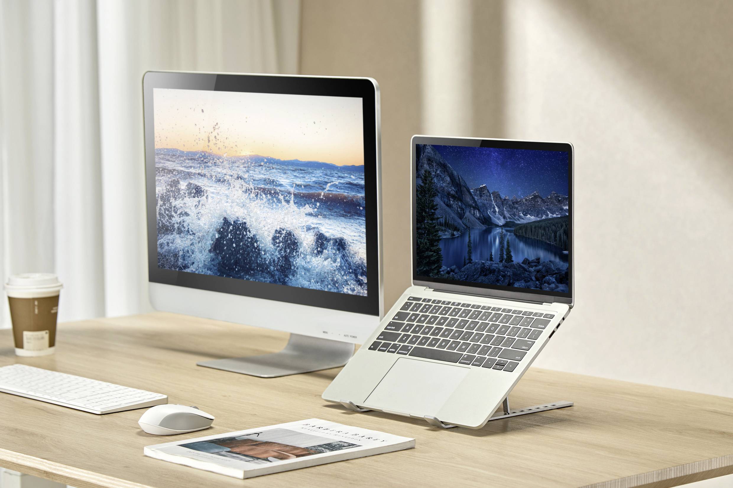 Stolní monitor a notebook zobrazující scénické přírodní obrazy na dřevěném stole; šálek kávy, klávesnice, myš a kniha poblíž.