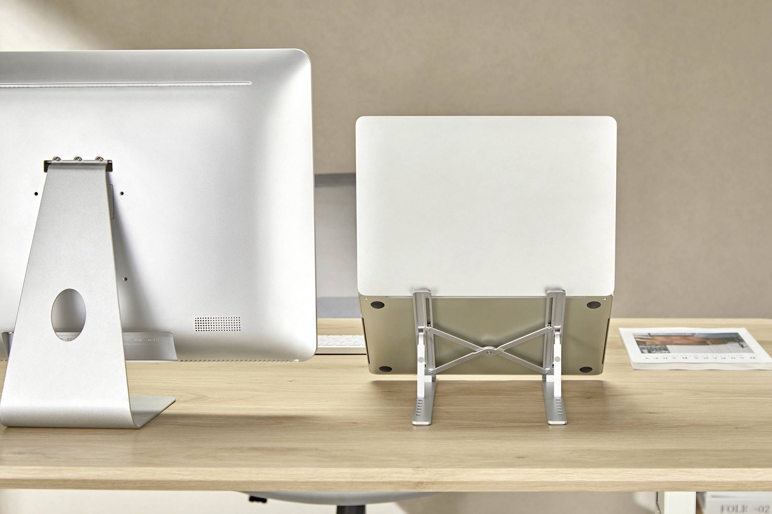 Minimalistický stůl s počítačovým monitorem, laptopem na stojanu a zarámovanou fotografií na pravé straně, umístěný na neutrálním pozadí.