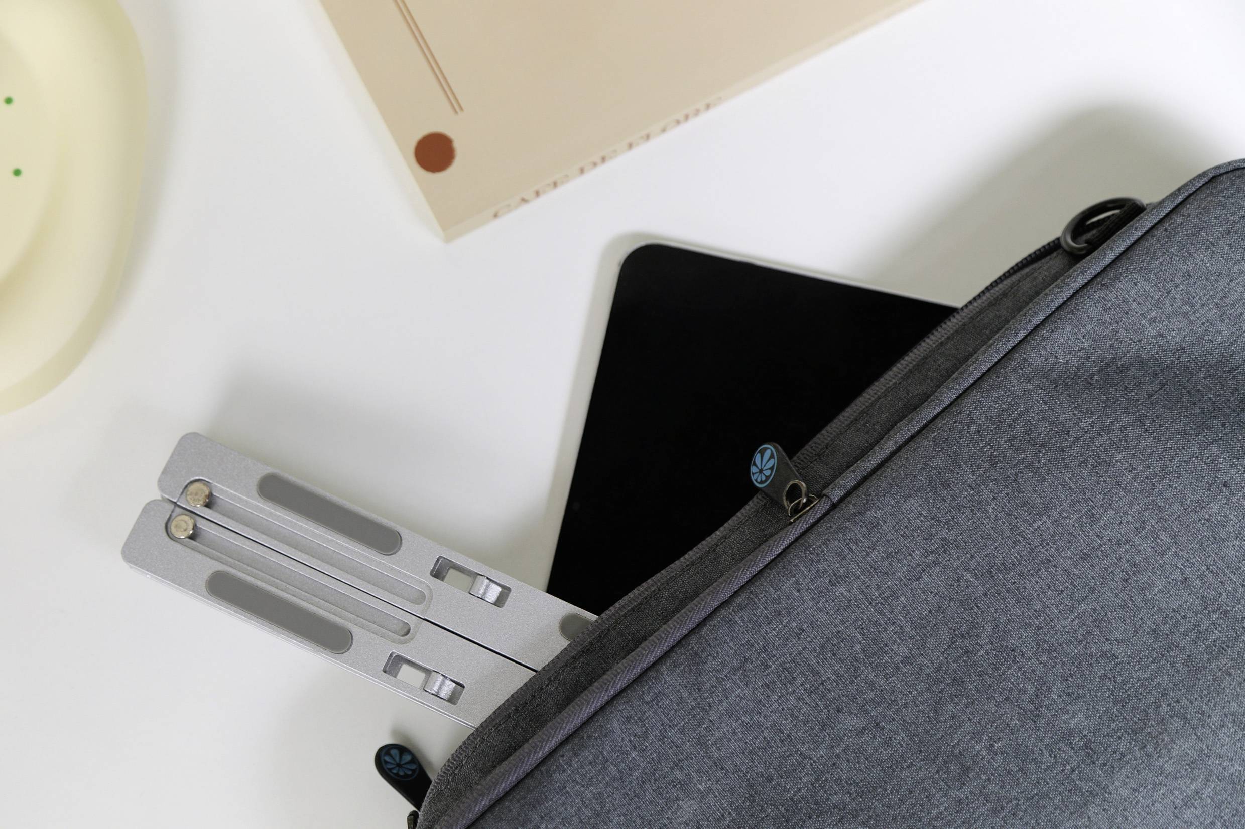 Šedý obal na notebook z části otevřený, odhalující tablet a skládací kovový stojan na notebook uvnitř, umístěný na bílém povrchu.