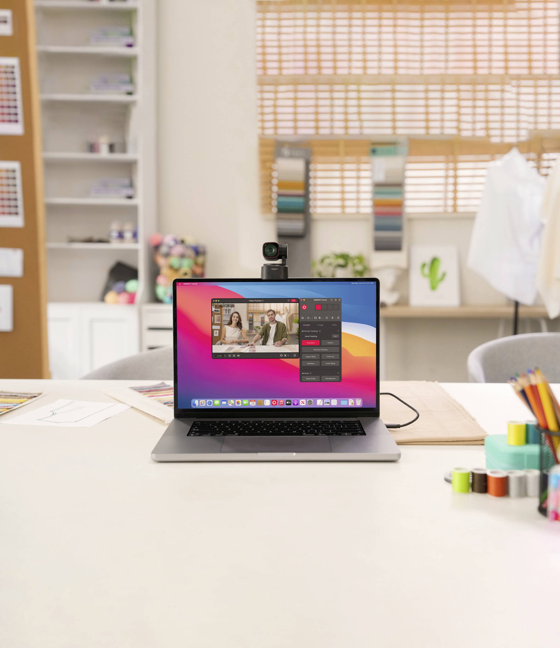 Laptop na stole zobrazuje videokonferenci. V pozadí jsou barevné vzorky a kancelářské potřeby.