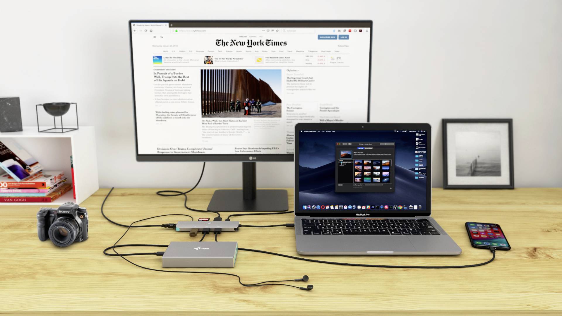 Psací stůl s monitorem zobrazujícím 'The New York Times', laptopem, fotoaparátem, smartphonem a připojenými kabely.