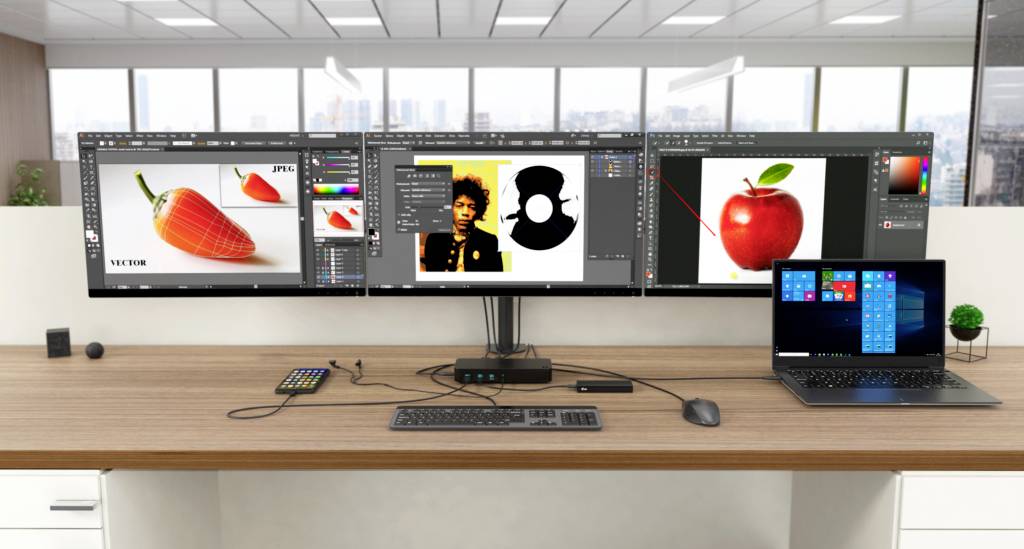 Psací stůl se třemi monitory a notebookem, který zobrazuje software pro úpravu obrázků a plochu systému Windows. V pozadí kancelářské vybavení.
