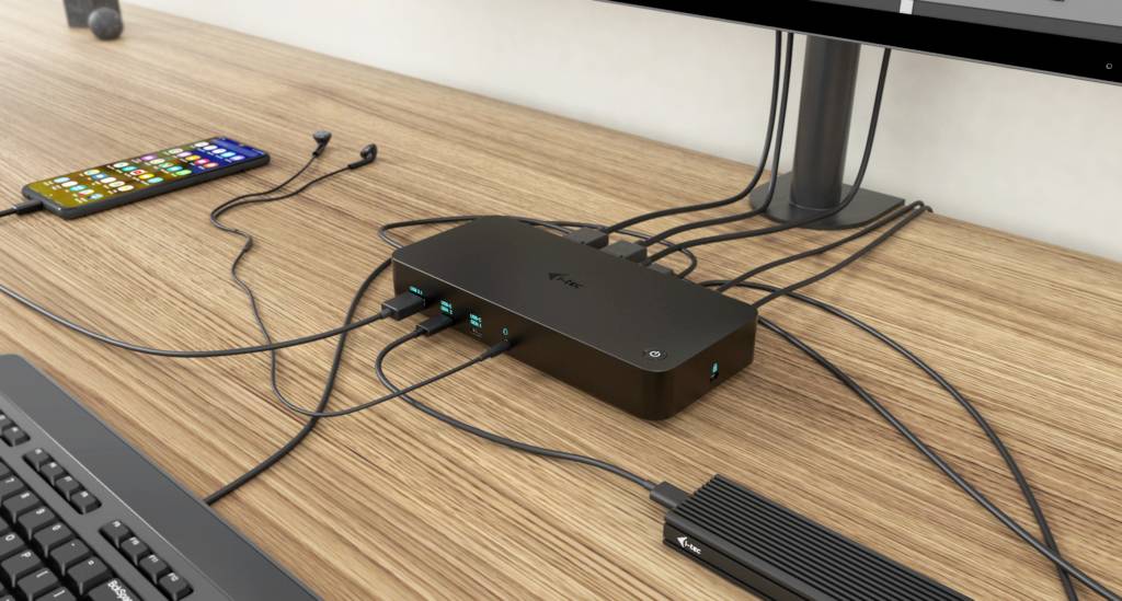 USB dokovací stanice s několika připojenými kabely na pracovním stole. Propojuje laptop, smartphone a periferní zařízení.