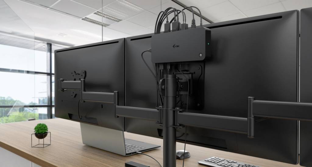 Dva počítačové monitory na psacím stole s malým zeleným květináčem, v moderní kanceláři s okny v pozadí.