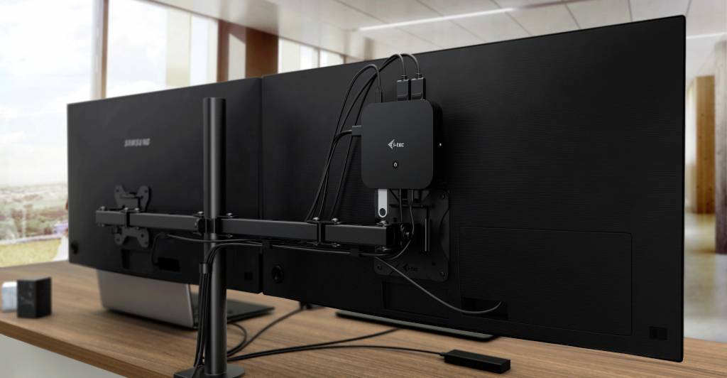 Dva velké počítačové monitory stojí vedle sebe na pracovním stole, oba upevněné na rameni monitoru. Vedení kabelů je patrné.