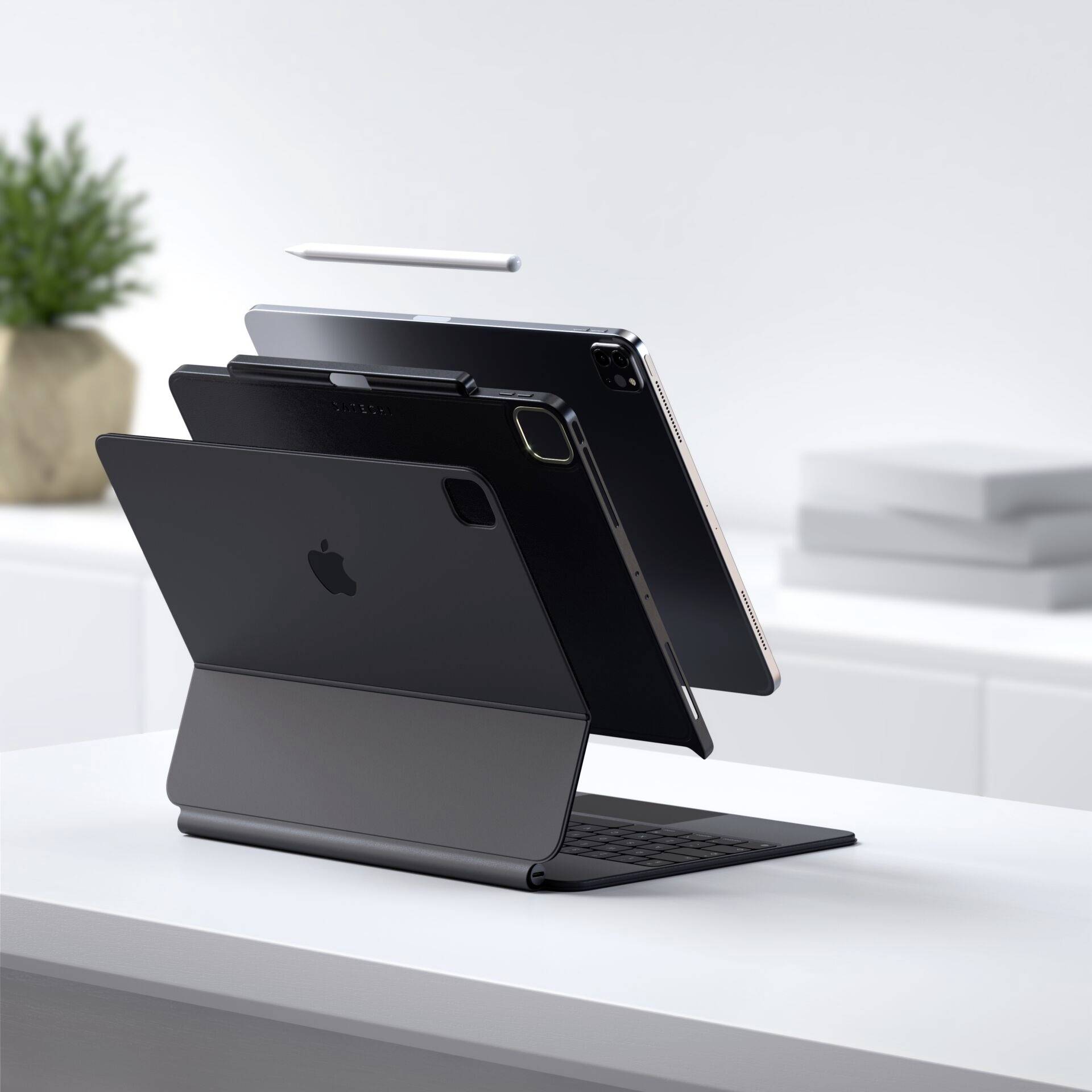 Tablet leží na krytu klávesnice, vedle něj volně visí pero. V pozadí jsou viditelné knihy a rostlina.
