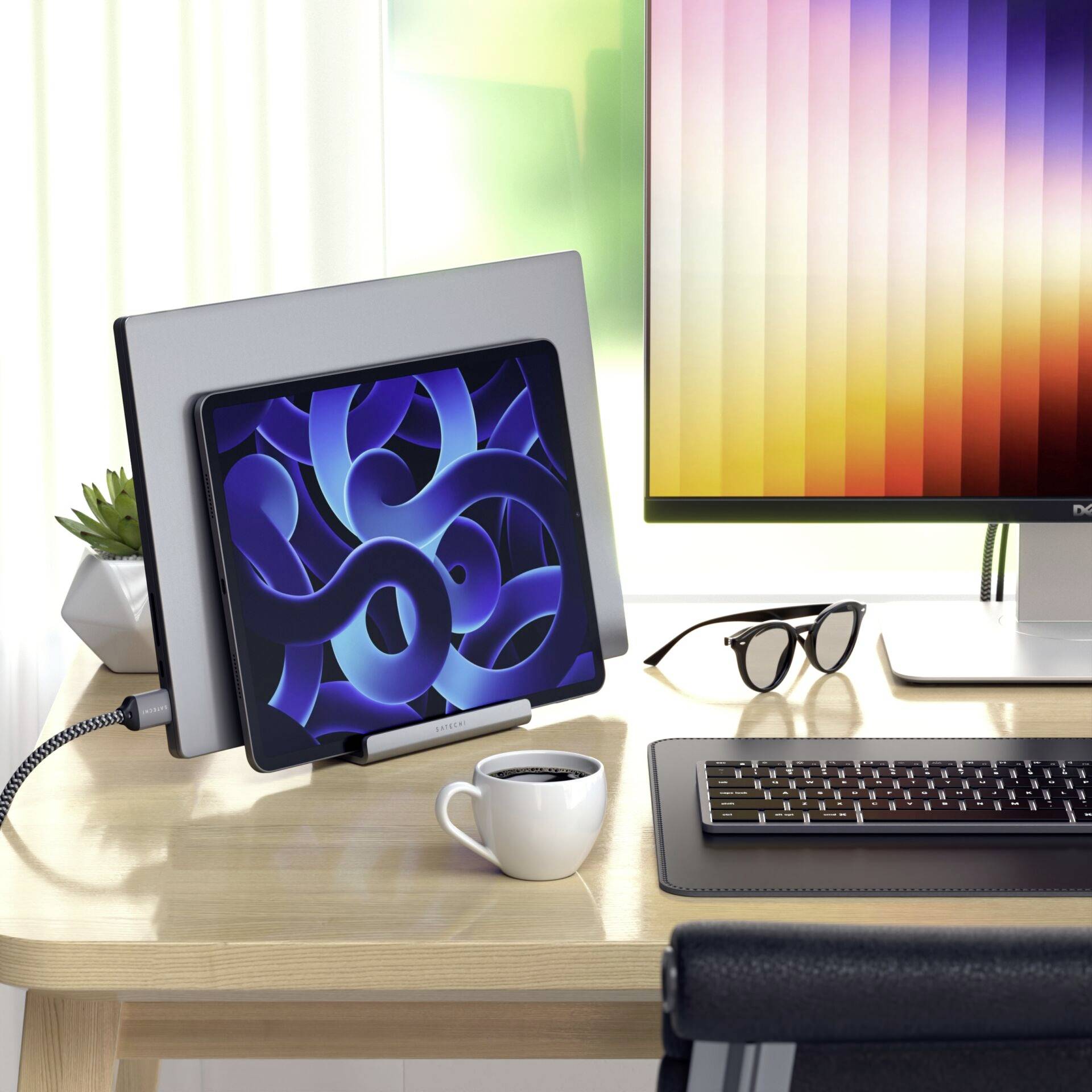 Kancelářský stůl s tabletem, monitorem, klávesnicí, brýlemi a šálkem. Monitor zobrazuje pestré vertikální pruhy, tablet abstraktní modré tvary.