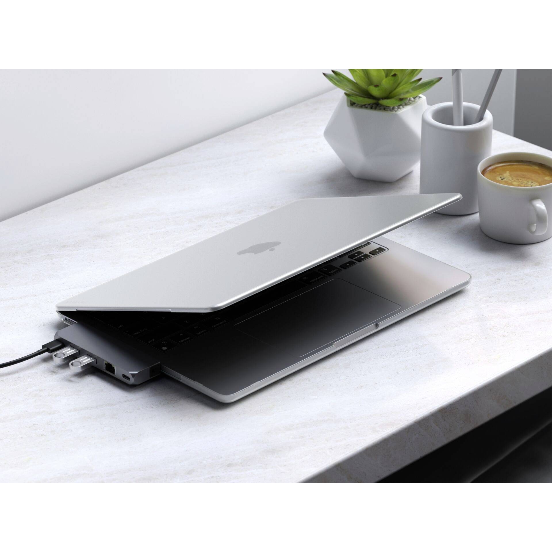 Částečně otevřený laptop s připojenými kabely na stole vedle šálku kávy a malé rostliny.