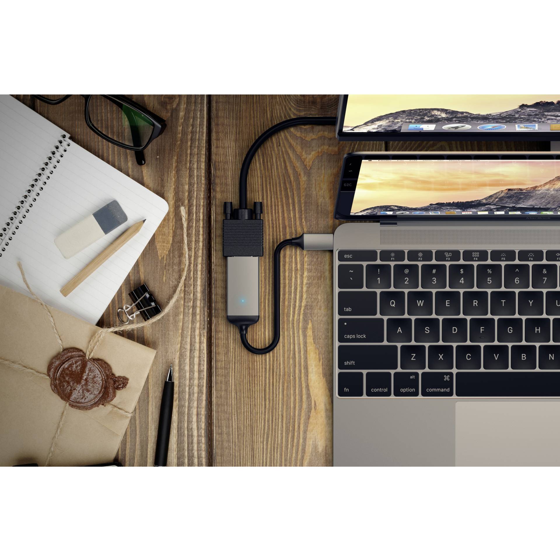 Laptop na dřevěném stole, připojený k USB rozbočovači. Vedle leží poznámkový blok, brýle, guma a zapečetěná obálka.