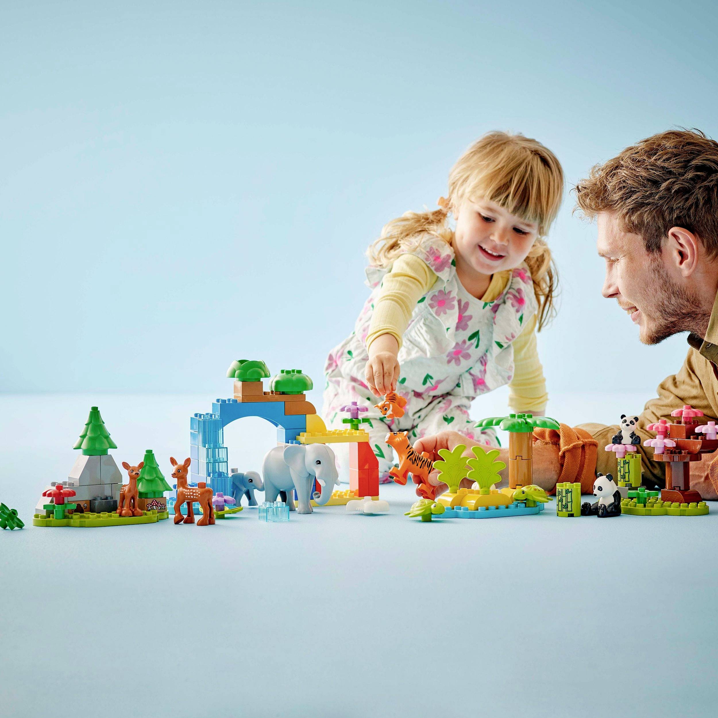 Dívka a muž si hrají s barevnými plastovými kostkami Lego a figurkami zvířat, které jsou uspořádány na stole.
