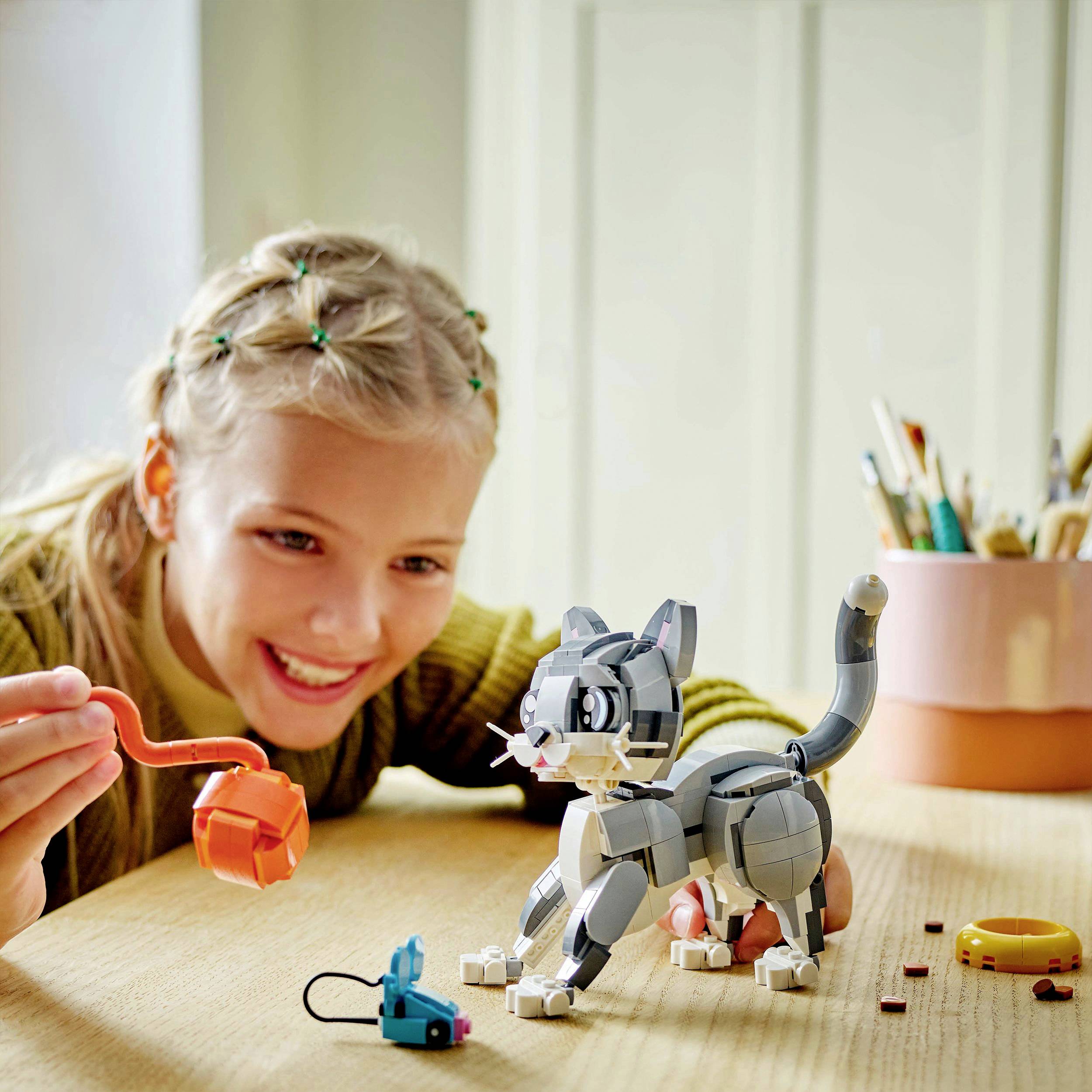 Dítě radostně hraje si s hračkou LEGO ve tvaru kočky na stole a přitom drží oranžové udičky.