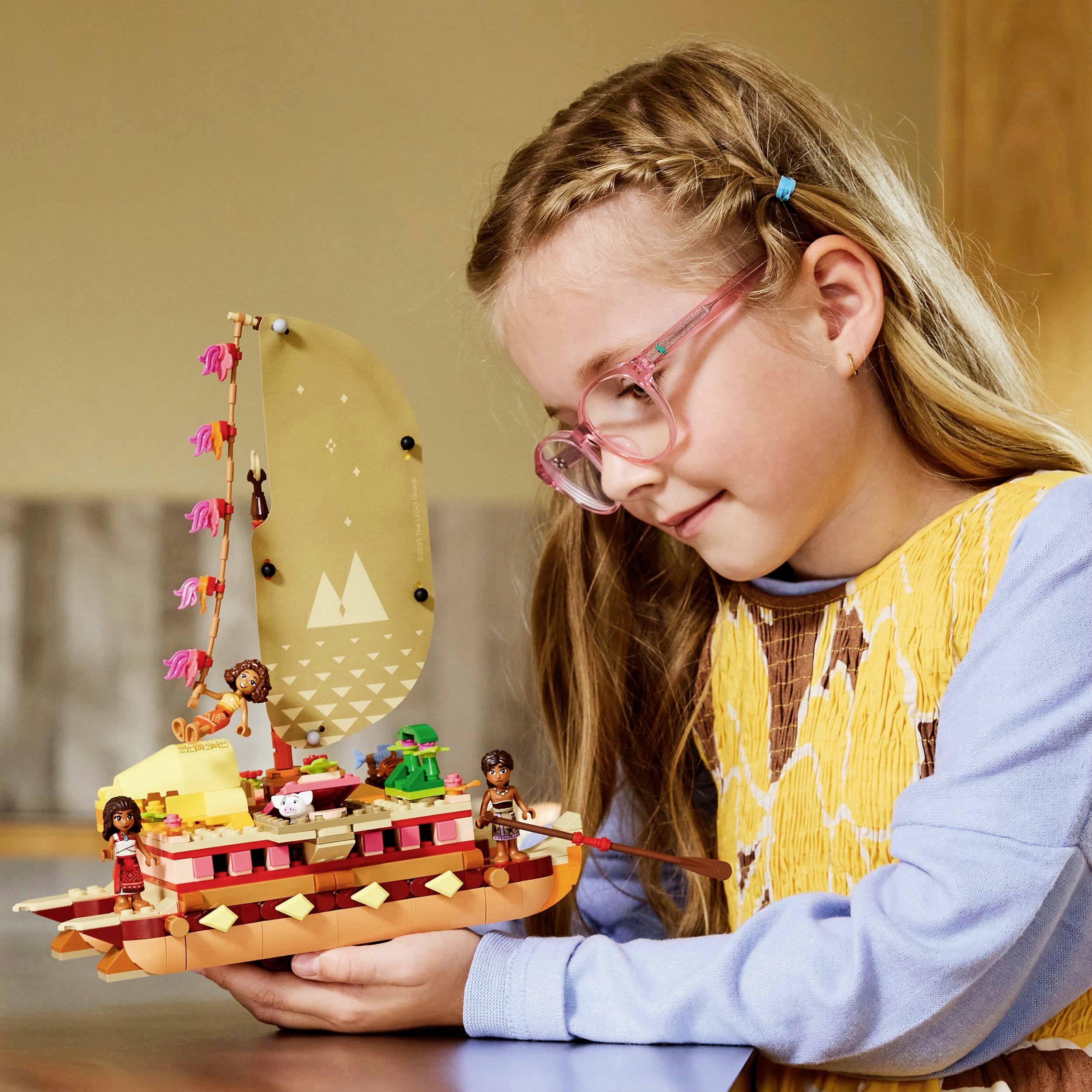 Dítě s brýlemi si hraje s barevnou LEGO lodí, která obsahuje figurky a plachtu. V pozadí je rozmazaný prostor.