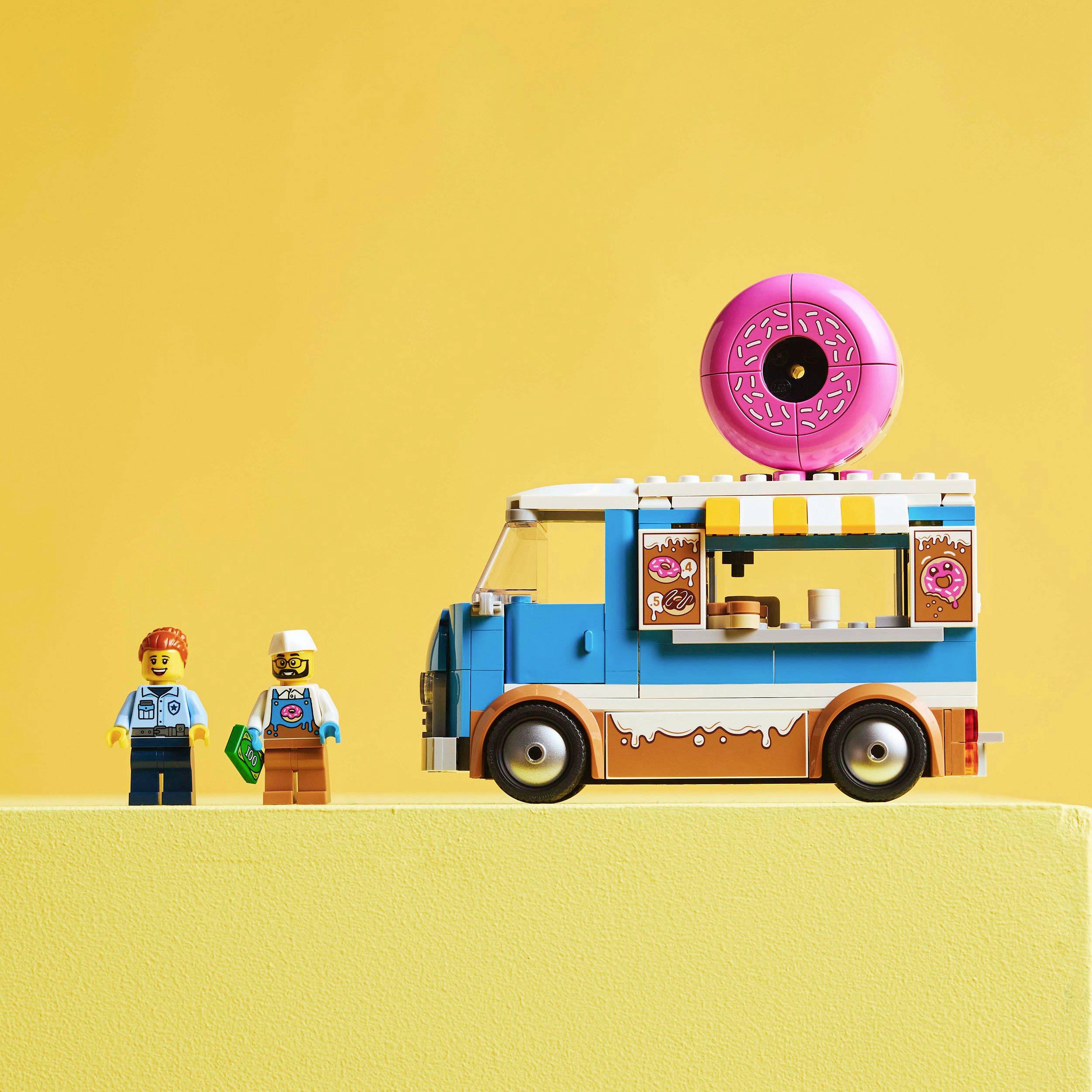 Malý model modrého food trucku na donuty s růžovým donutem na střeše a dvěma figurkami vedle něj na žlutém pozadí.