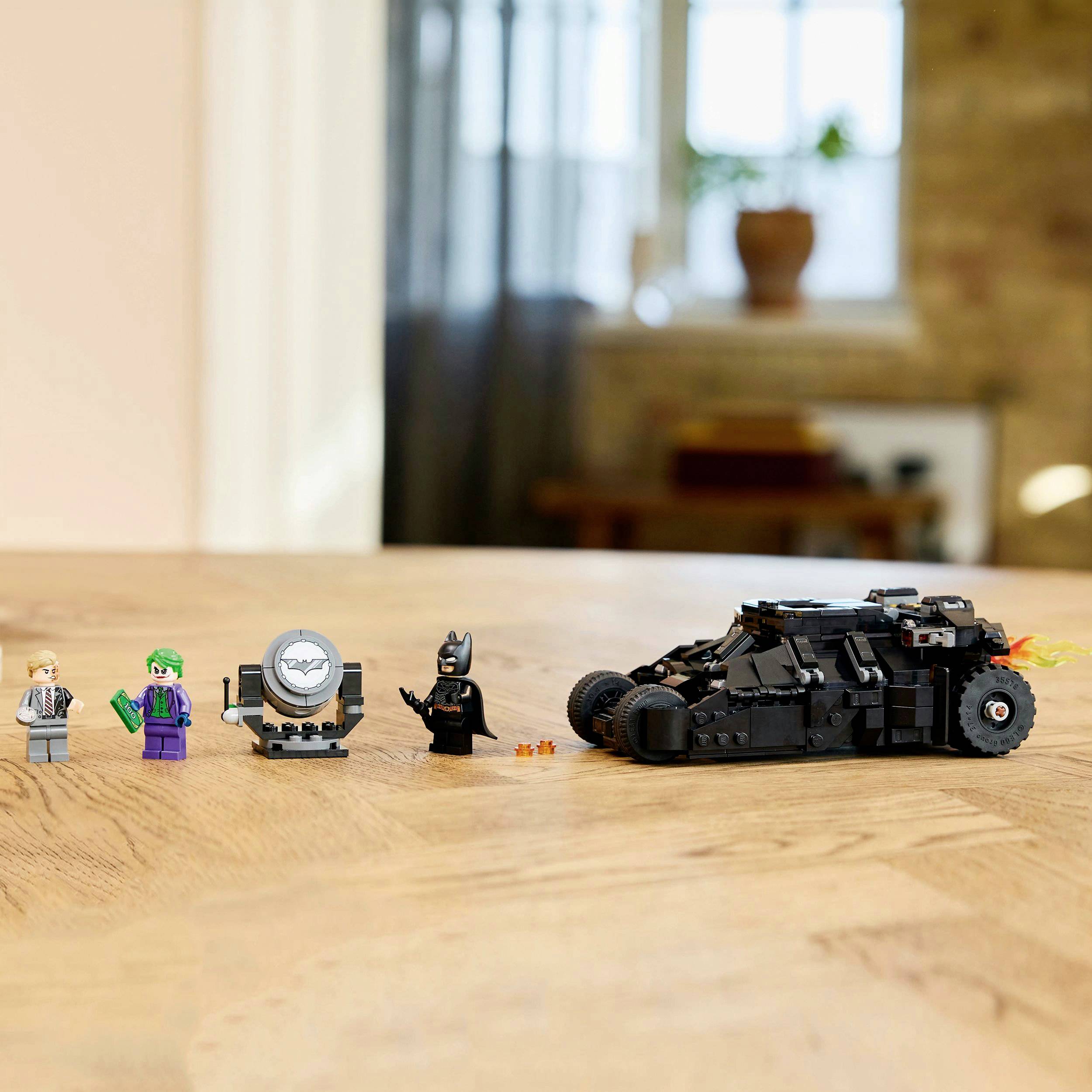 Lego scéna s Batman figurkami, Jokerem, Bat-signálem a Batmobilem na dřevěném stole, rozmazané pozadí.