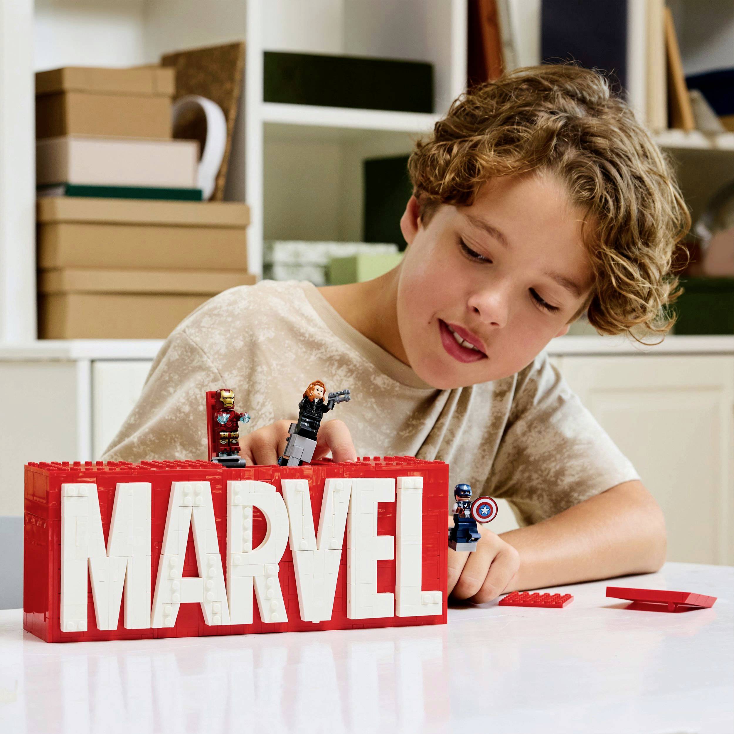 Dítě si hraje s LEGO a staví písmena 'MARVEL' z červených kostek. Vedle toho stojí minifigurky superhrdinů na stole.