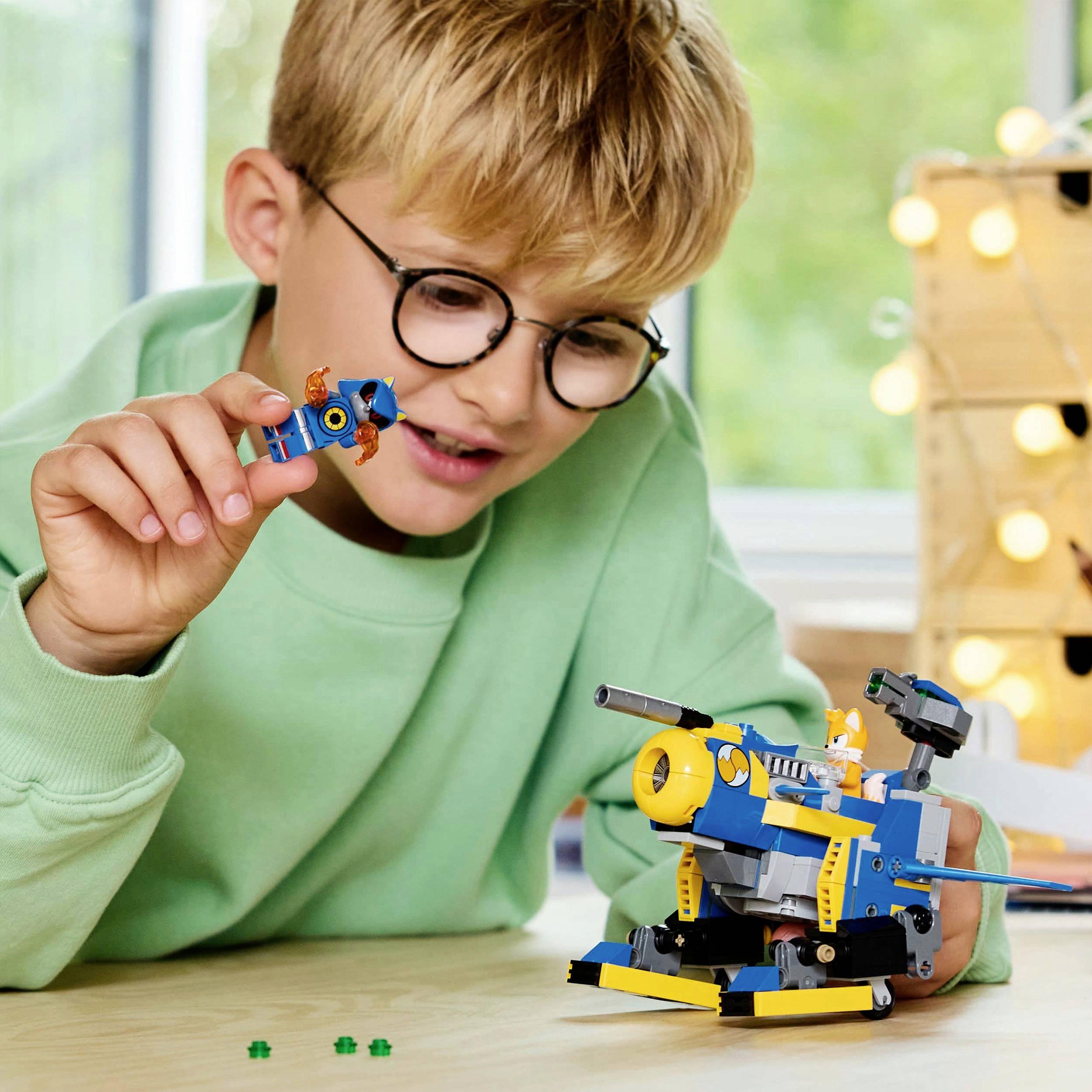 Dítě staví fotoaparát z Lego kostek, je patrná jeho soustředěnost a kreativita. V pozadí jsou světla a okna.