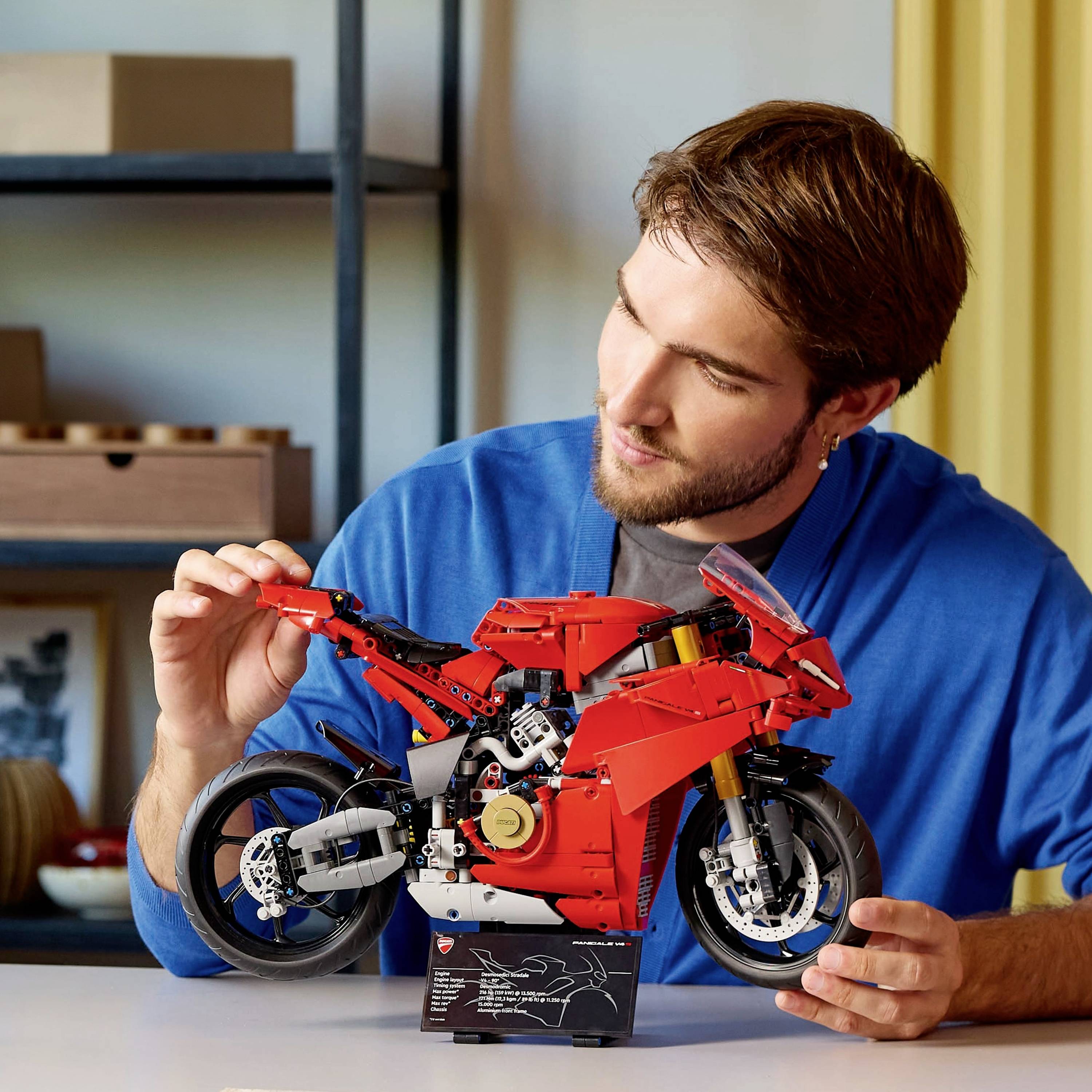 Muž v modrém svetru montuje detailní, červený model motocyklu ze stavebních dílků a soustředěně se na něj dívá.