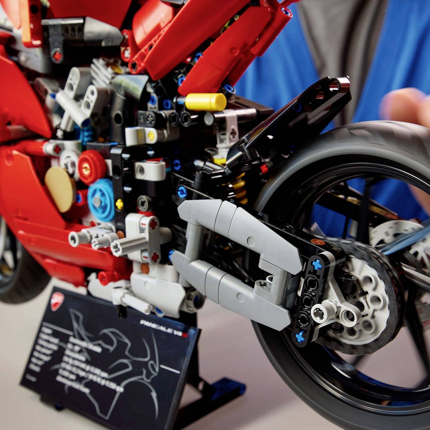 Detailní model červené motorky ze stavebních dílků, který zobrazuje zadní kolo a motorový prostor, v pozadí rozmazaná postava.