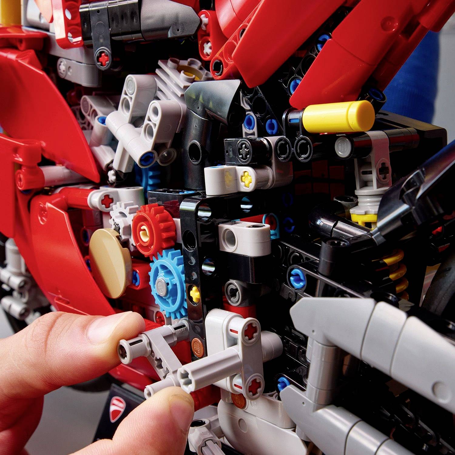 Osoba staví komplexní robotický model z barevných dílků Lego Technic. Model ukazuje ozubená kola a pohyblivé části.