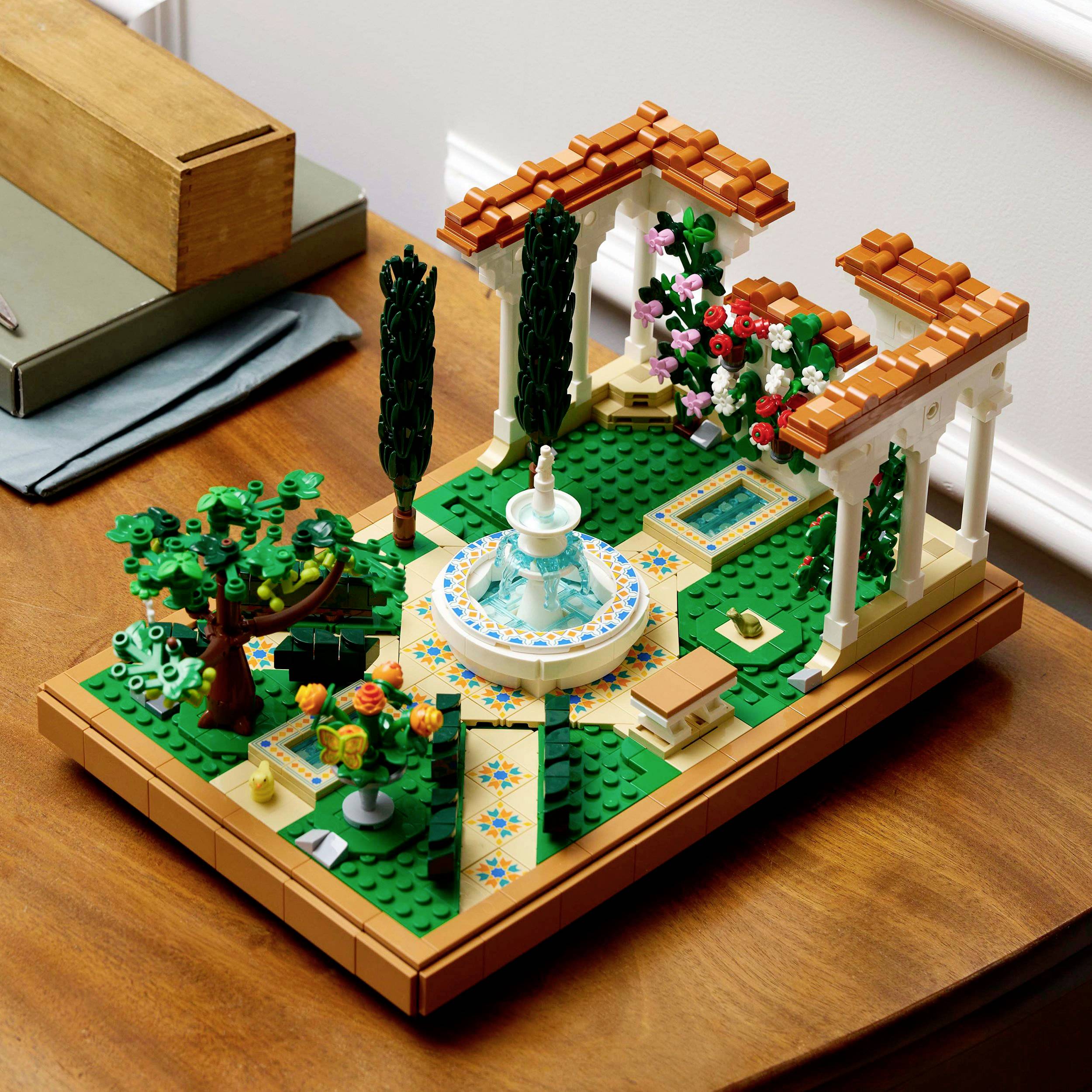 Model LEGO zahrady s centrální fontánou, obklopené stromy, květinami a sloupy na dřevěném stole.