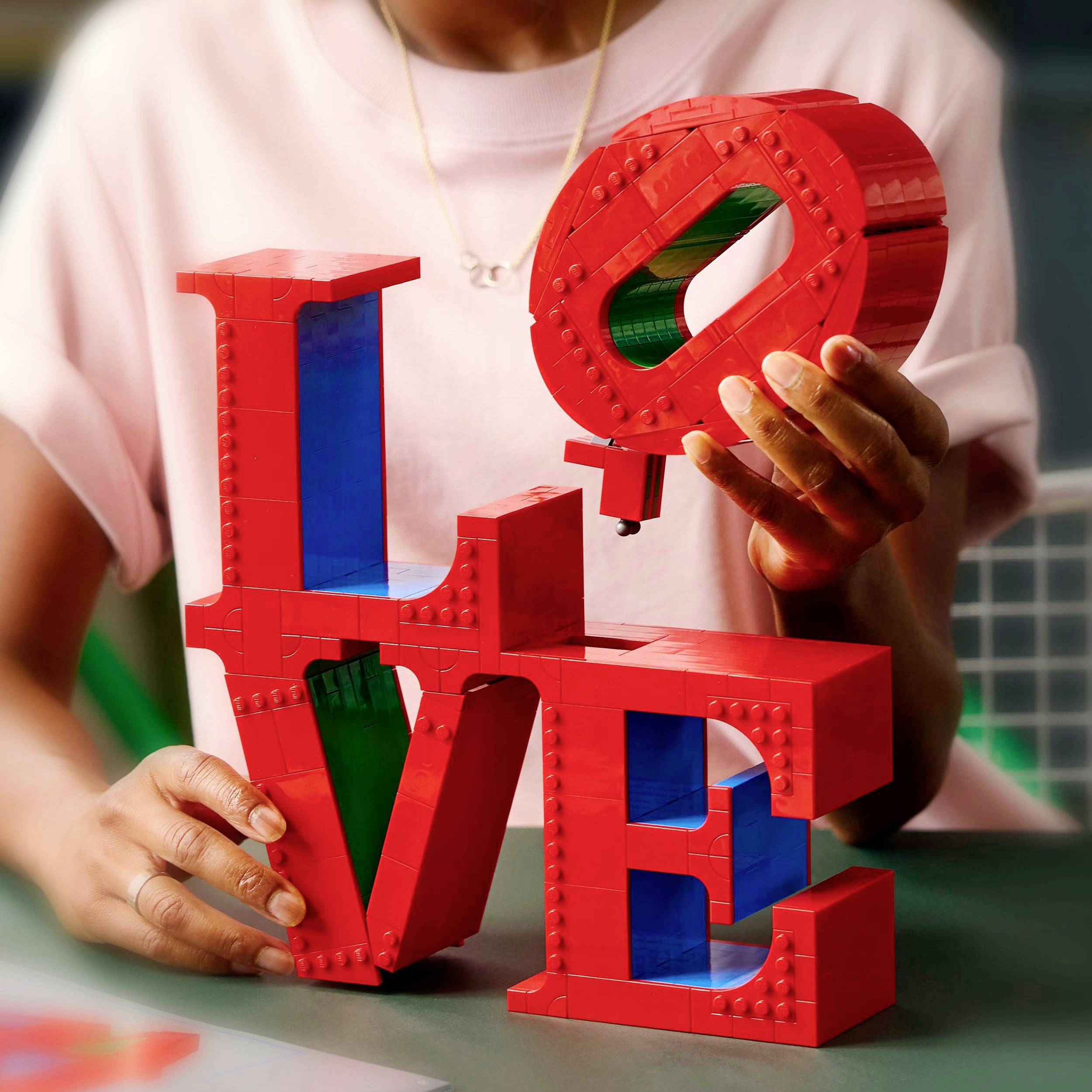 Osoba drží barevný LEGO model slova 'LOVE'; ukazuje kreativní způsob stavby s červenými a modrými díly.