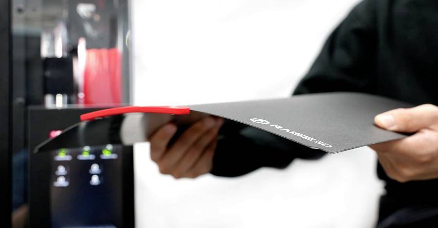 Osoba drží flexibilní 3D tiskovou podložku před 3D tiskárnou. Podložka se ohýbá, aby umožnila snadné odstranění červeného vytištěného objektu.