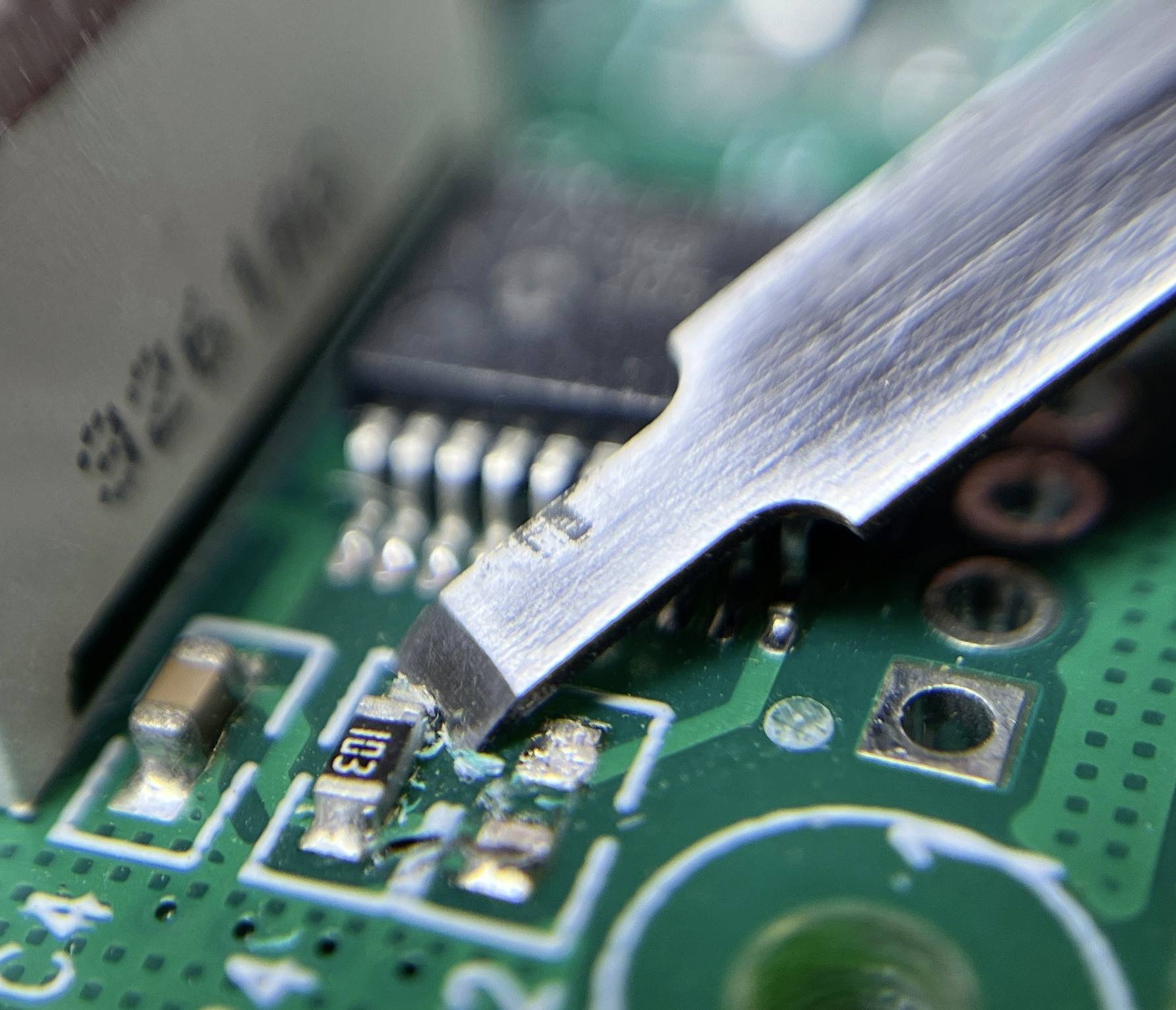 Detailní záběr pájecí pistole opravující zelenou desku plošných spojů, zobrazující elektronické komponenty a pájené spoje na povrchu.