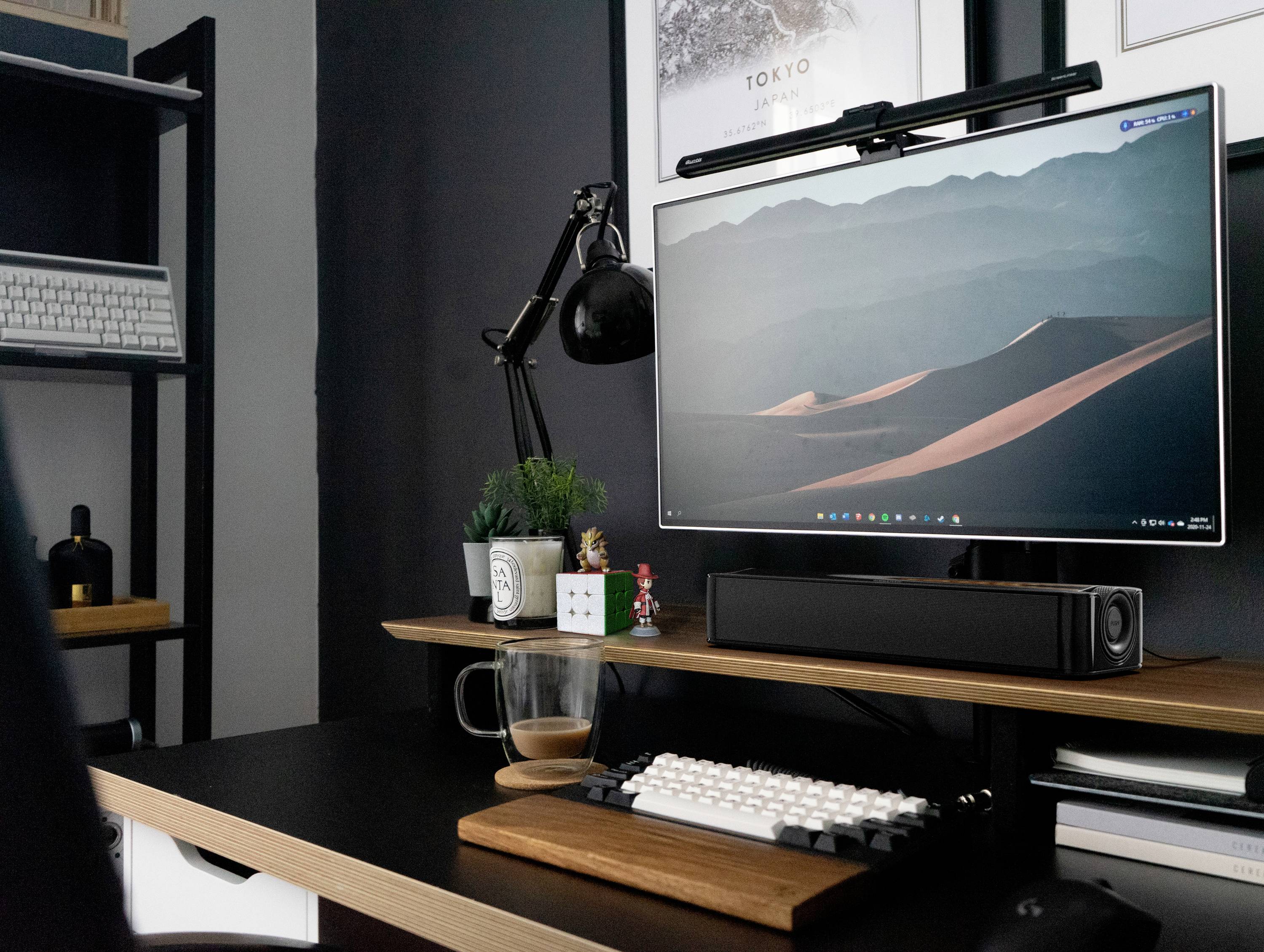 Moderní psací stůl s monitorem zobrazujícím pouštní krajinu. Klávesnice, lampa, reproduktory a rostliny jsou uspořádány pro kreativní práci.