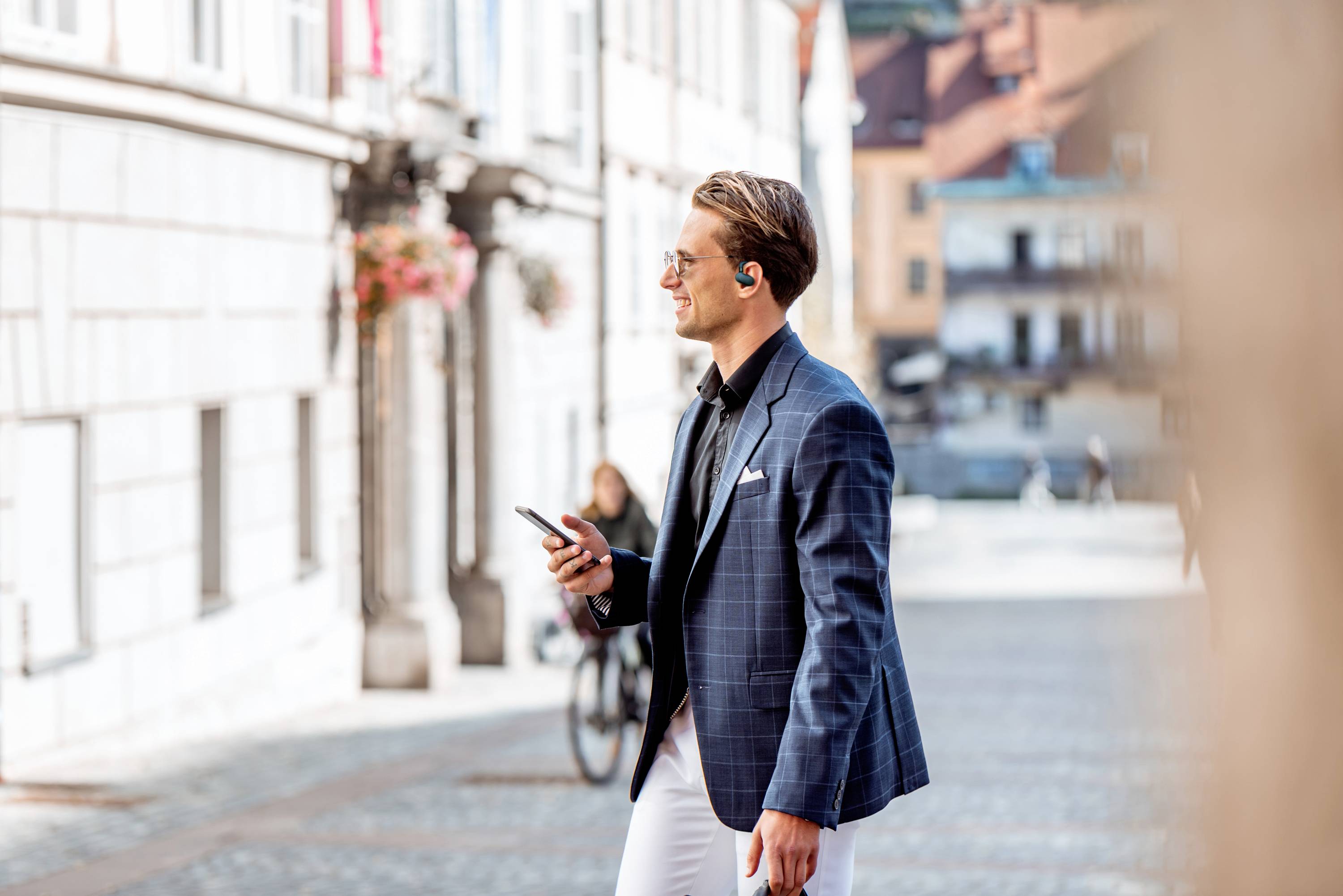 Muž v světle modrém obleku kráčí po slunné ulici a dívá se na svůj smartphone. V pozadí jede cyklista.