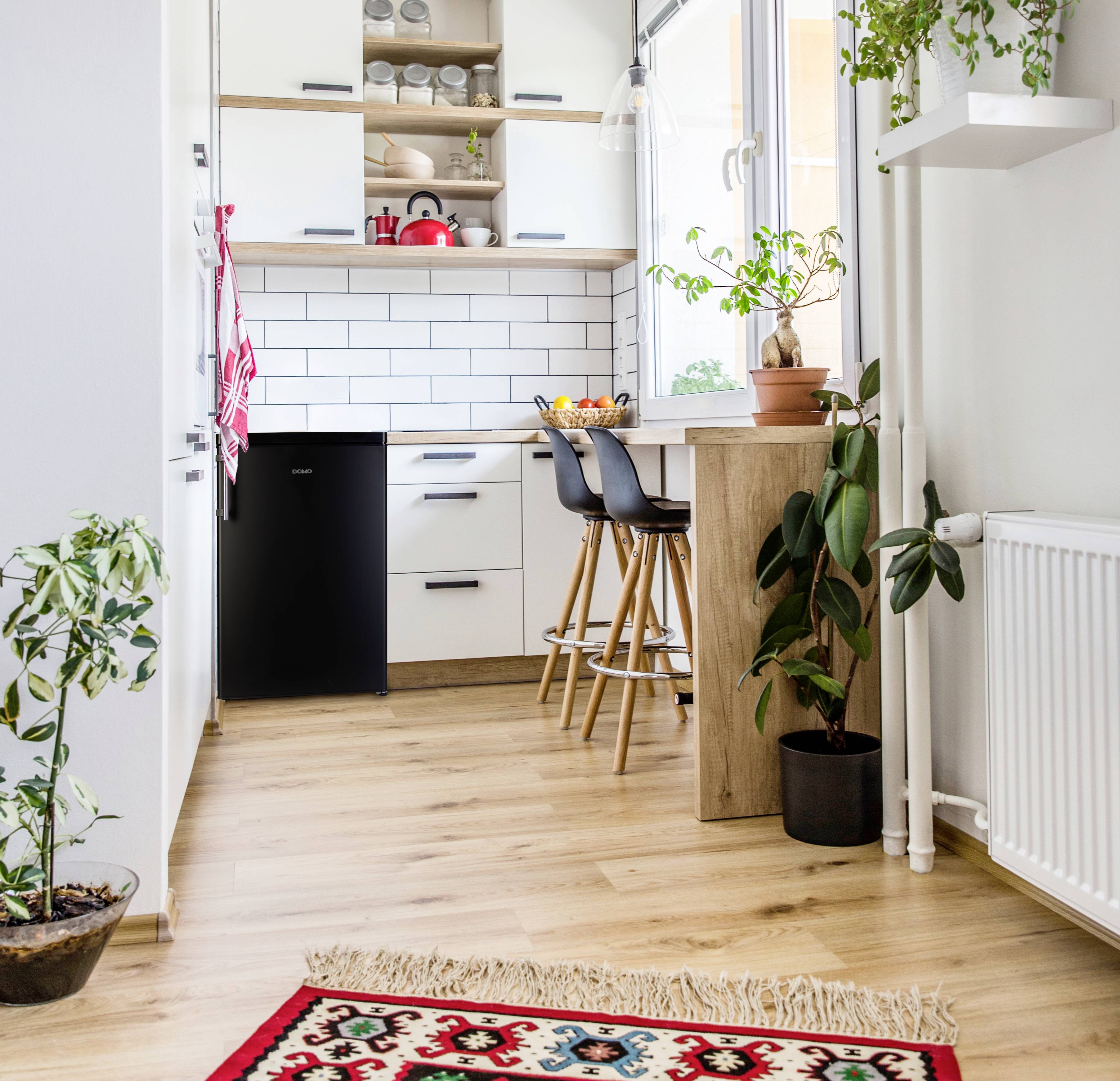 Moderní, světlá kuchyně s dřevěnou podlahou a bílými skříňkami. Rostliny stojí na podlaze a na pultu, který má dvě barové stoličky.
