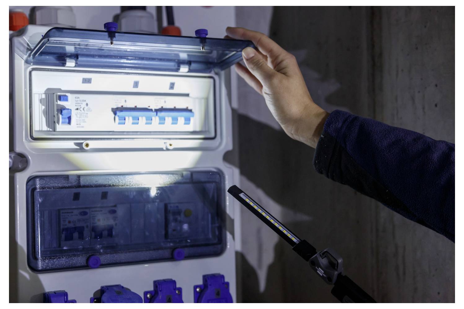 Osoba kontrolující otevřený elektrický rozvaděč s osvětlenými jističi, přitom drží dveře, což naznačuje údržbářské nebo instalační práce.