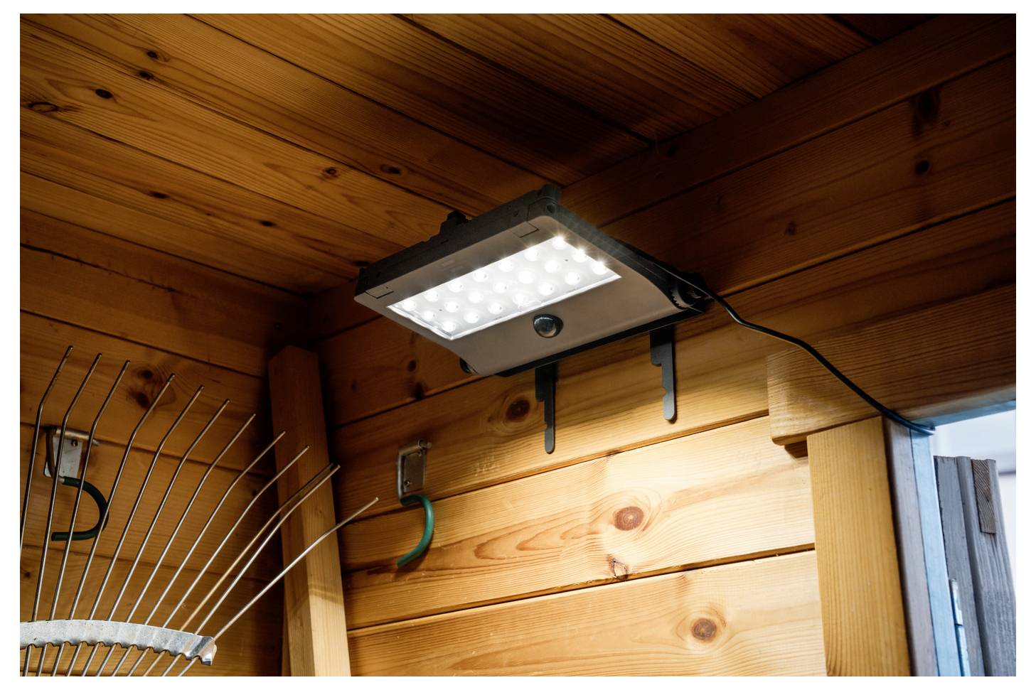 LED bezpečnostní světlo namontované pod dřevěným přesahem střechy, osvětlující zeď a oblast dveří venku.