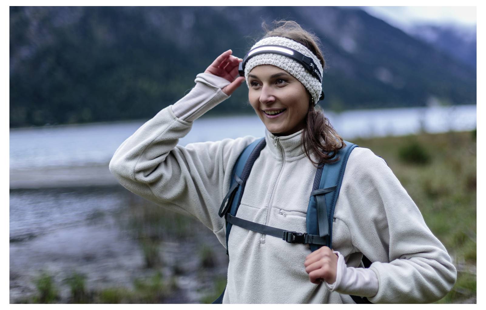 Usměvavá žena v outdoorovém oblečení si upravuje čelenku během túry poblíž jezera s horami v pozadí.