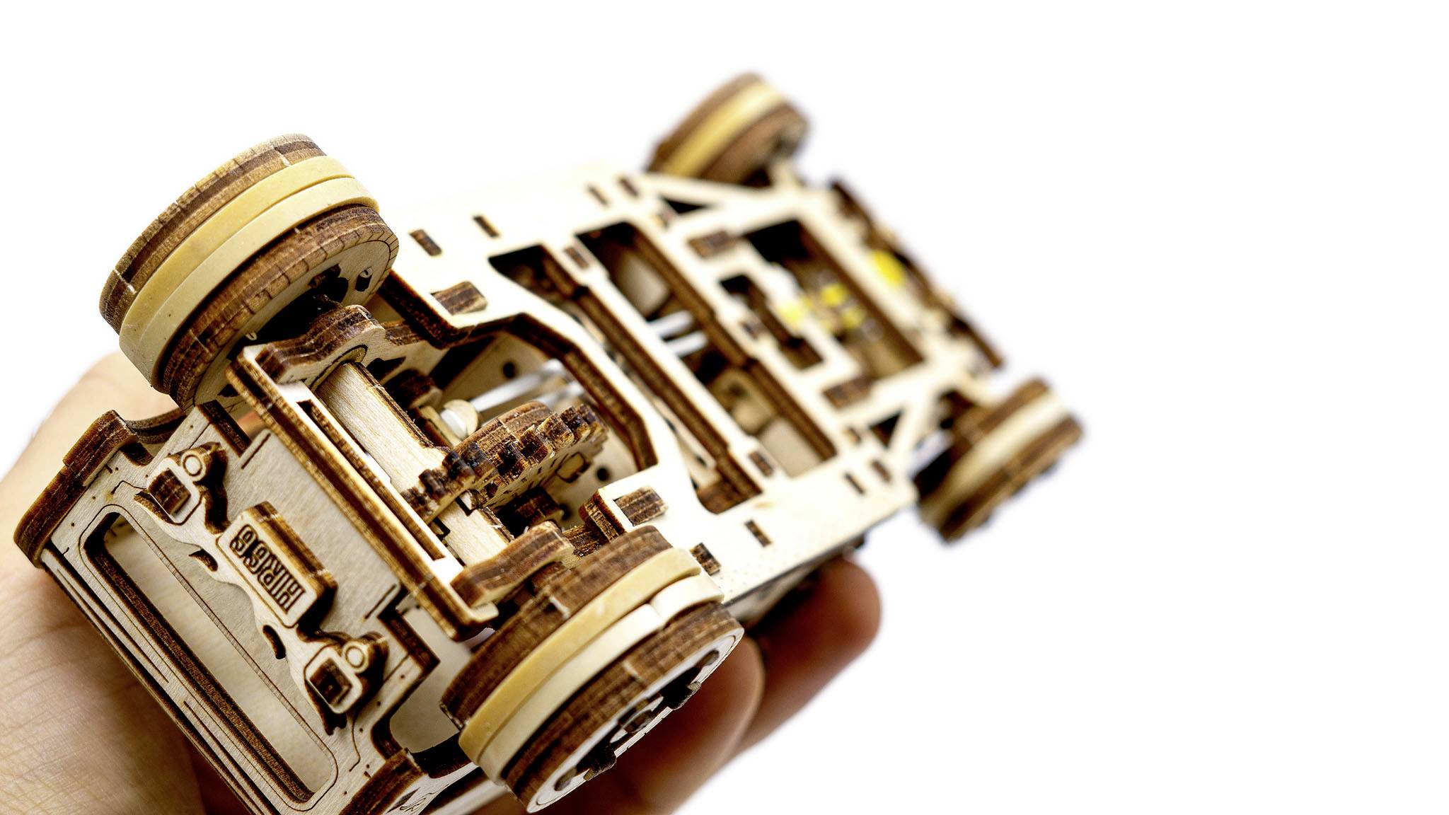 Jedna ruka drží detailní dřevěný model auta s komplexními mechanickými strukturami na bílém pozadí.