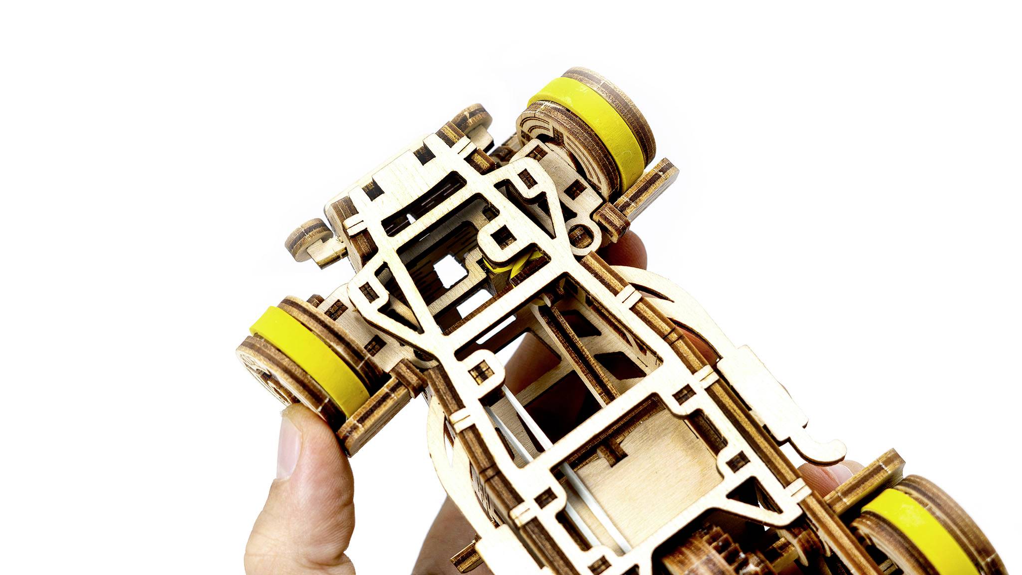 Jedna ruka drží dřevěný model automobilu se žlutými koly, který poukazuje na složité mechanické struktury.