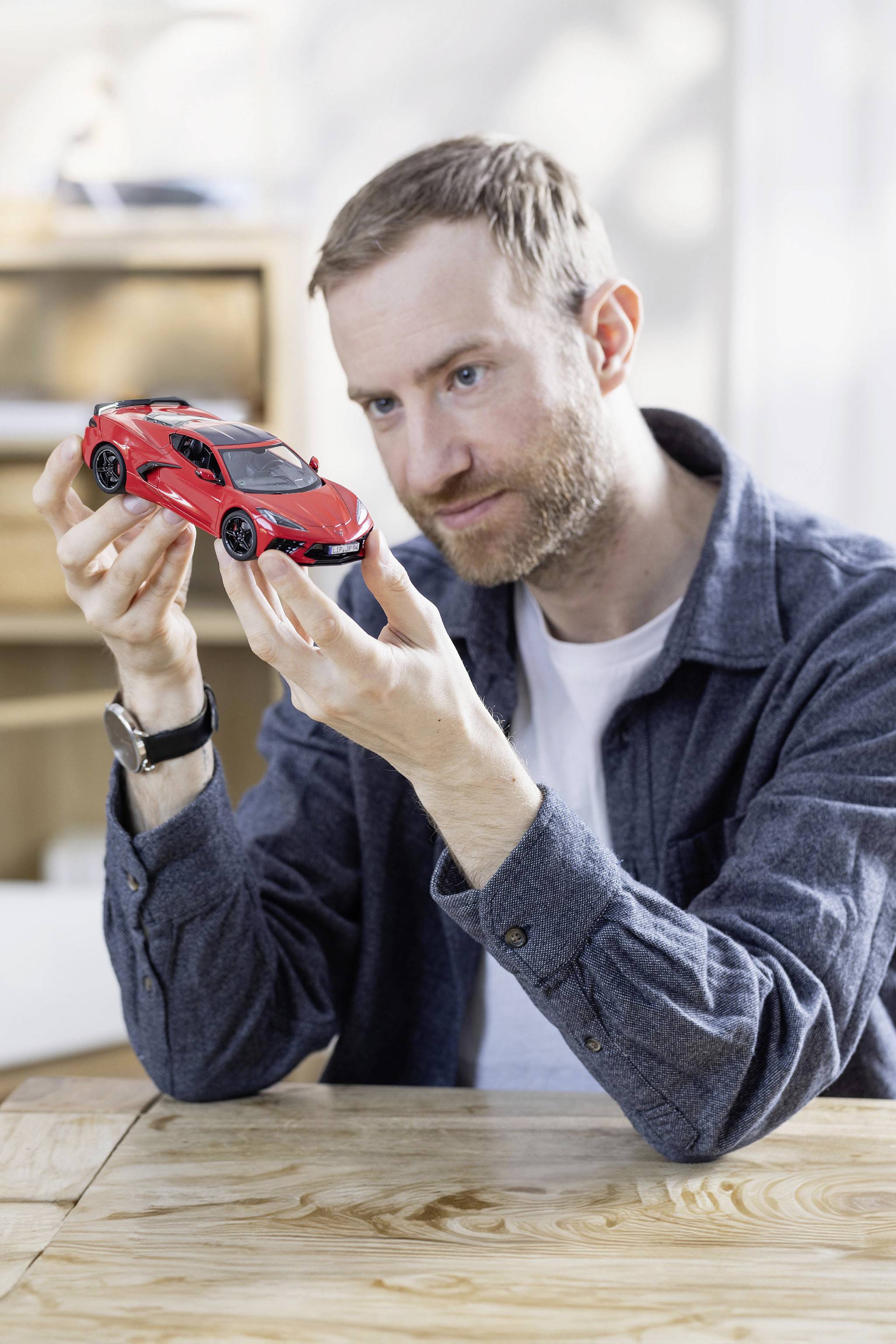 Muž drží a prohlíží si modelové auto ve tvaru červeného sportovního vozu na dřevěném stole.
