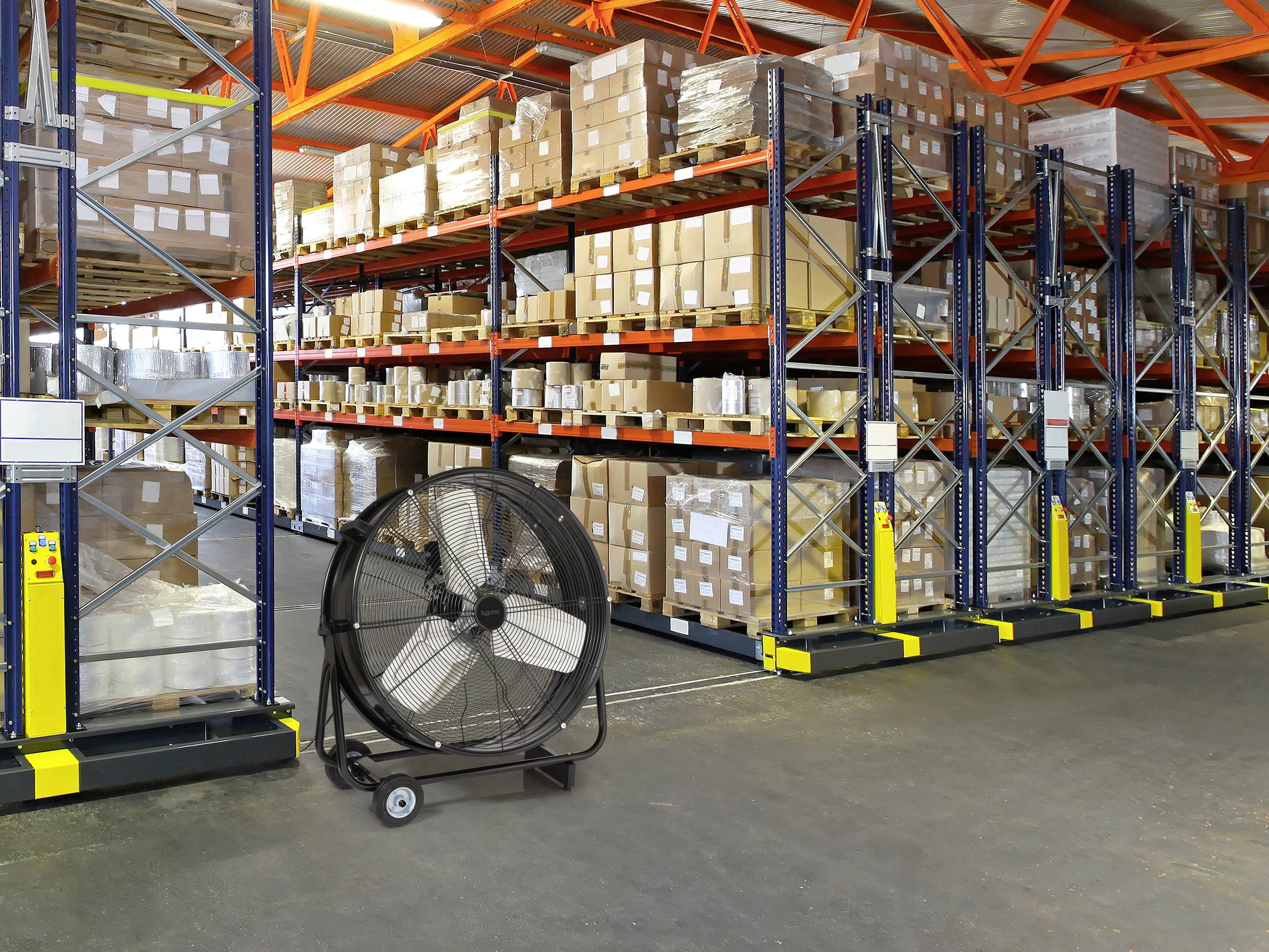 Ve skladu stojí regály s naskládanými krabicemi. Na podlaze v popředí se nachází velký pojízdný ventilátor.