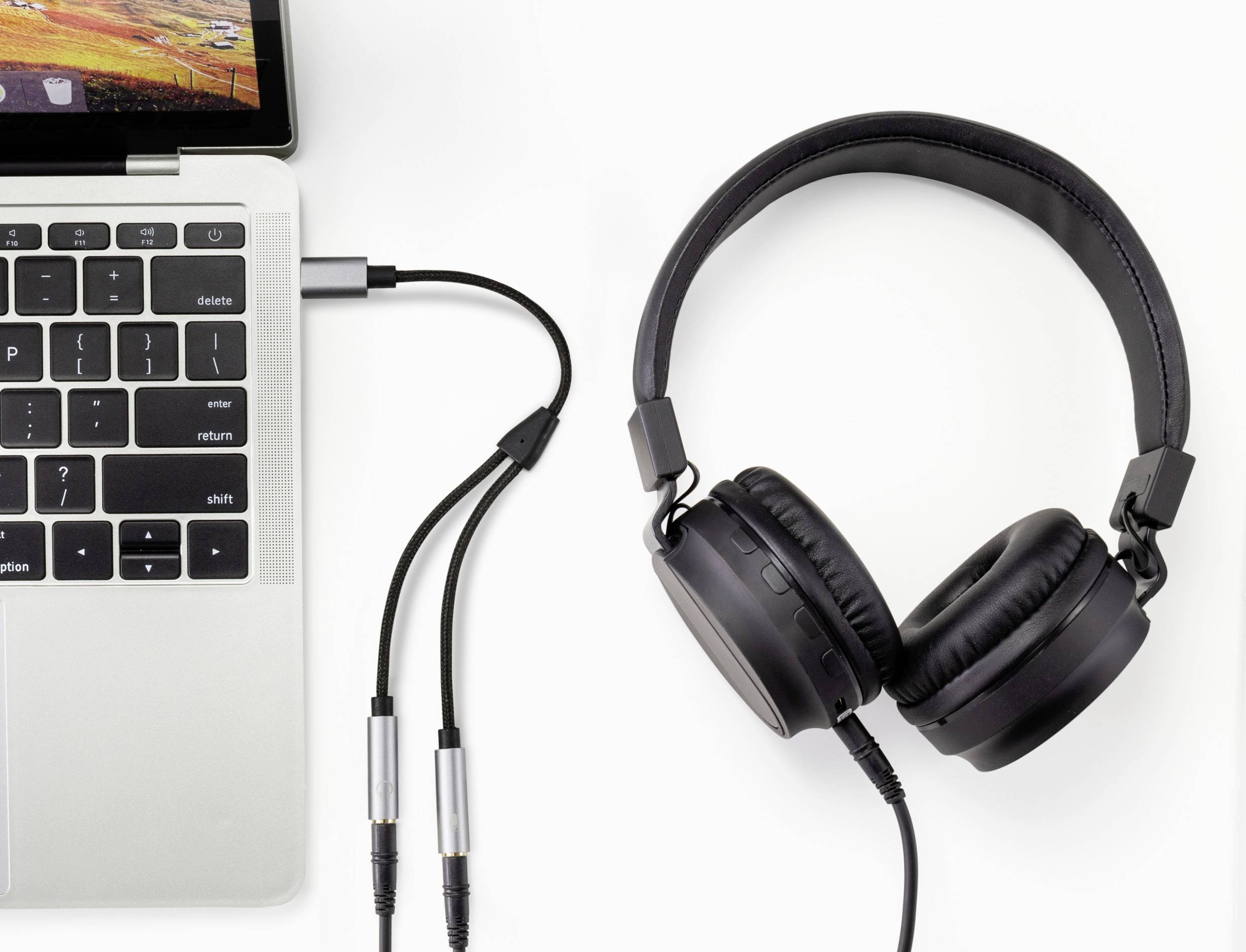 Laptop s připojenými sluchátky prostřednictvím audio kabelu. Laptop je umístěn v levém horním rohu bílého pozadí.