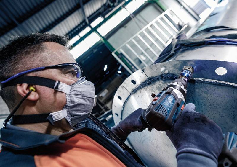 Dělník v ochranných brýlích a respirátoru používá vrtačku při práci na velkém kovovém konstrukčním dílu.