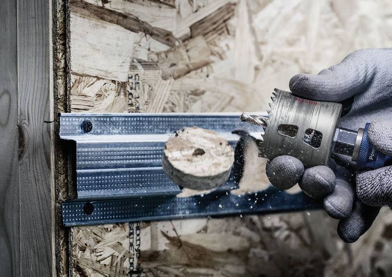 Ruka drží vrtačku s kotoučovou pilou, která vrtá díru do dřevěné desky. Na desce je viditelný kovový nosník.