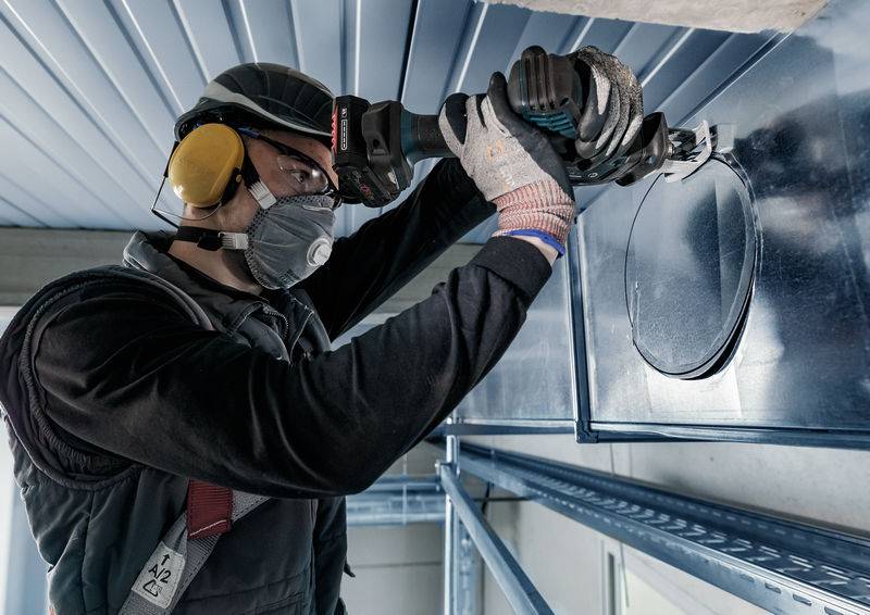 Dělník v helmě, masce a chráničích sluchu používá nástroj k vystřižení kulatého otvoru do ventilačního systému.