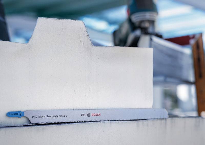 Pilový list s nápisem 'PRO Metal Sandwich precise BOSCH' přesně prořezává bílý materiál, v pozadí jsou viditelné nástroje.