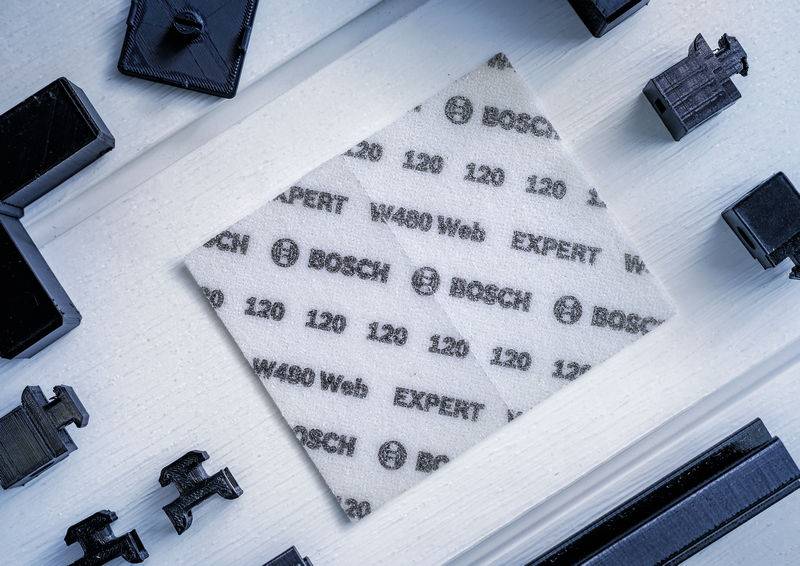Brusný papír s potiskem 'Bosch Expert' uprostřed, obklopený černými geometrickými plastovými díly na bílém pozadí.